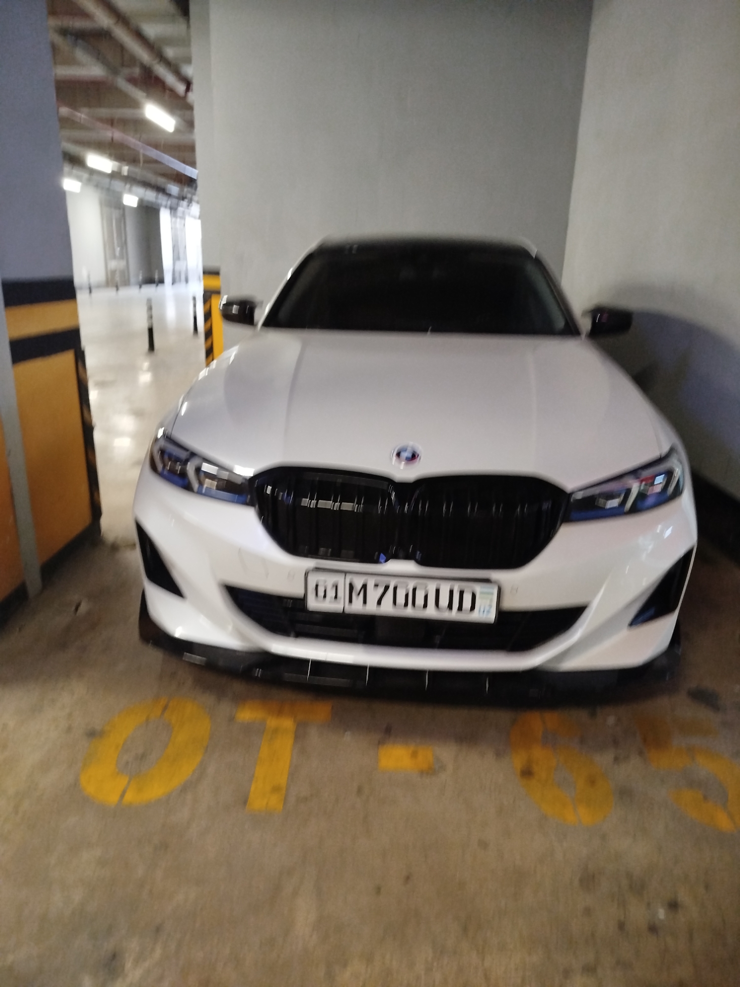 A white BMW sedan is parked in a concrete parking garage in Tashkent, Uzbekistan. The car is facing the camera, and its headlights are illuminated. The license plate of the car reads "01 M700UD". The parking garage has yellow and black striped pillars and overhead lighting. The floor of the garage is concrete and has yellow markings, some of which are partially visible and appear to be numbers.