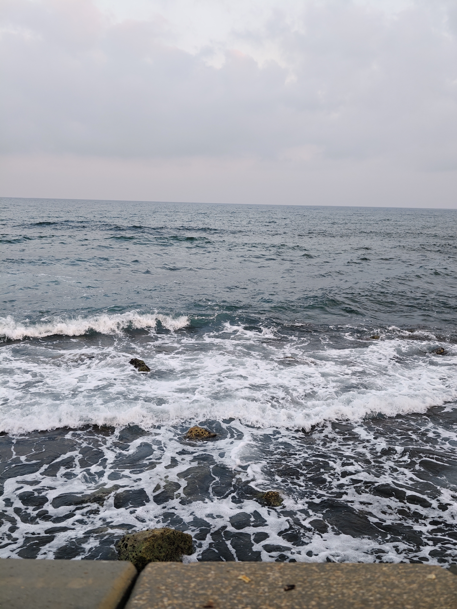 This image depicts a view of the Mediterranean Sea from the coast of Tel Aviv, Israel. The sea is characterized by moderate waves crashing onto a rocky shoreline, creating white foam that churns around dark, submerged rocks. The water itself appears a deep, muted blue-gray, reflecting the overcast sky. The sky is filled with a thick layer of gray clouds, obscuring any direct sunlight and contributing to the subdued lighting of the scene. In the foreground, a concrete structure, likely part of a breakwater or promenade, is visible, with its textured surface and weathered appearance. The overall atmosphere is calm and somewhat somber, typical of a cloudy day at the beach. There are no people visible in the frame.