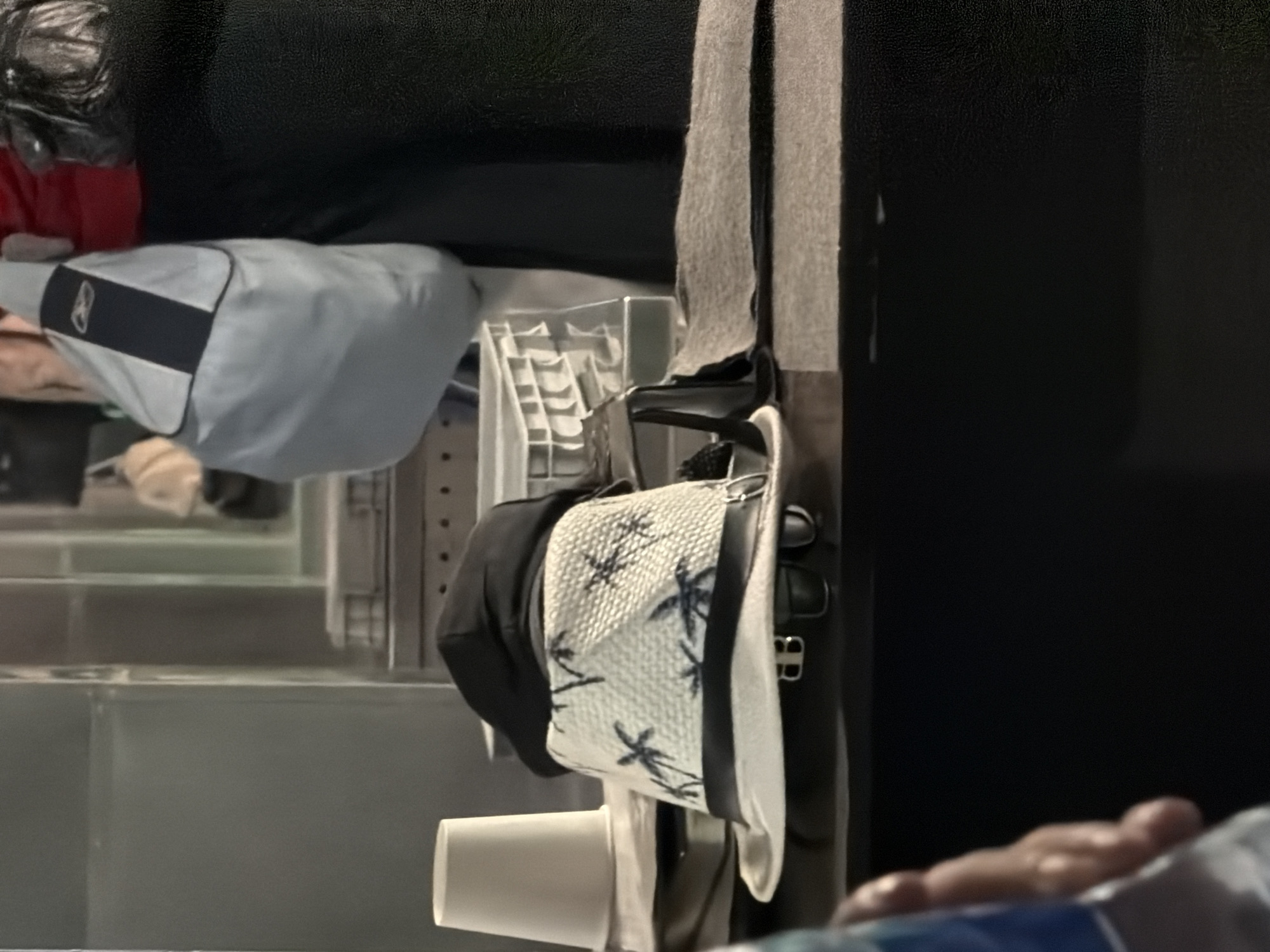 An indoor scene shows a white paper cup to the left, partially obscuring a stacked set of white plastic containers in the background. In the foreground, a white fedora with a black band and a pattern of dark palm trees sits on a surface, topped by a black cap. Below the fedora, a pair of sunglasses is visible. To the right, a section of a dark textured surface is present, with a light-colored fabric swatch resting on it. In the far background, a light gray backpack with a dark stripe is visible, along with red and black garments. A person's arm and hand are partially visible in the bottom left corner of the frame.