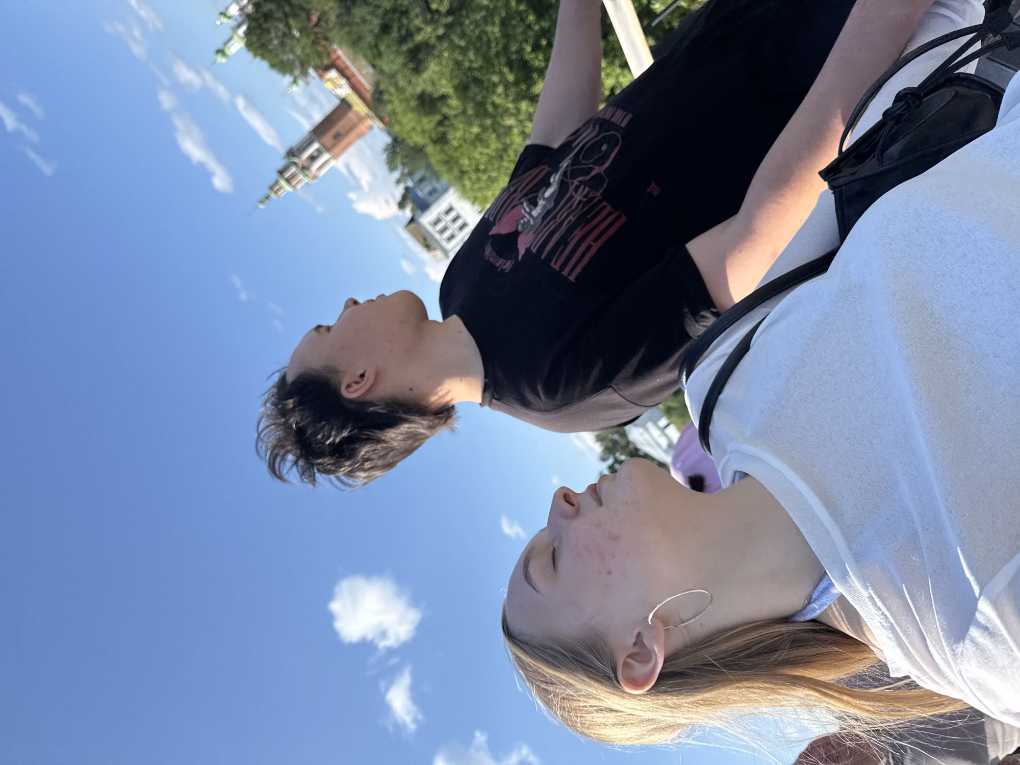 In Cherdyn, Russia, two individuals are captured in a candid outdoor shot against a bright blue sky dotted with white clouds. A young woman with blonde hair is positioned in the foreground, her eyes closed and face tilted upwards, appearing to bask in the sunlight. She wears a white t-shirt and a large silver hoop earring. Beside her, a young man with dark, styled hair is also looking upwards, his profile visible. He is dressed in a black t-shirt with a graphic print. In the background, a distinctive multi-tiered brick tower with green accents, characteristic of local architecture, rises above lush green trees. The overall scene suggests a moment of leisure and appreciation for the surroundings, likely taken on a clear, sunny day.