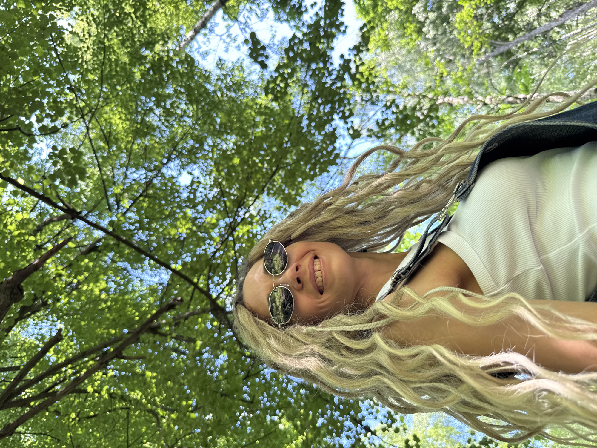 A woman with long, blonde dreadlocks and sunglasses smiles up at the camera from a low angle, positioned outdoors under a canopy of green trees and a blue sky. She is wearing a white tank top and denim overalls. The background is filled with dappled sunlight filtering through the leaves, creating a bright and airy atmosphere. The location is specified as Rusanovo, Russia.