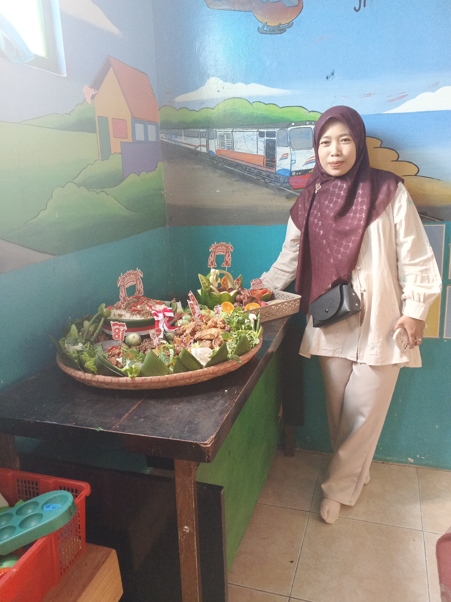 A woman in a maroon hijab and a light-colored tunic and pants stands to the right of a large, round woven tray filled with food. The tray is a centerpiece, decorated with small flags and what appears to be Indonesian food items like rice, fried chicken, and vegetables. The woman is holding a smartphone in her right hand and has a small black purse slung over her left shoulder. The background is a mural depicting a cartoon train and a house. The scene appears to be indoors, possibly in a classroom or community space, with a simple wooden table supporting the food tray and red plastic bins visible beneath it. The floor is tiled. The overall ambiance suggests a celebratory or communal gathering. The location is identified as Warungkiara, Indonesia.
