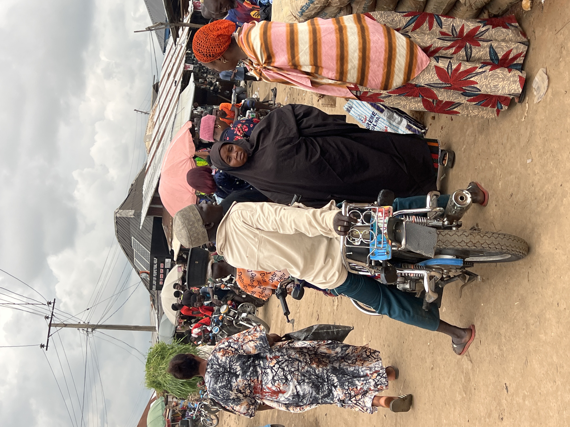 This media file depicts a bustling market scene in Asaba, Nigeria, likely during the daytime given the overcast sky. The image is oriented vertically, showing a lively gathering of people amidst stalls and various objects.
The central focus is a group of individuals, some interacting with a motorcycle. A person wearing a light-colored jacket and dark pants is seated on the motorcycle, while another person in a black headscarf and traditional attire is standing nearby. Further back, a woman in a striped pink and orange outfit and an orange head covering is visible, along with a pink umbrella. Other individuals are scattered throughout the scene, some carrying goods or browsing. The ground appears to be dusty, typical of an outdoor market.
Notable details include the presence of several motorcycles, suggesting a common mode of transportation or goods conveyance in this location. A portion of a building with "OUR LADY OF PERPETUAL HELP" and what appears to be religious imagery is visible in the background, hinting at the proximity of a church or religious institution. There are also signs with some text, including "DEBR" and "UREME OLINA," which are partially legible. The overall atmosphere seems busy and active, with a sense of community engagement. The cloudy sky suggests a potential for rain or diffused sunlight.