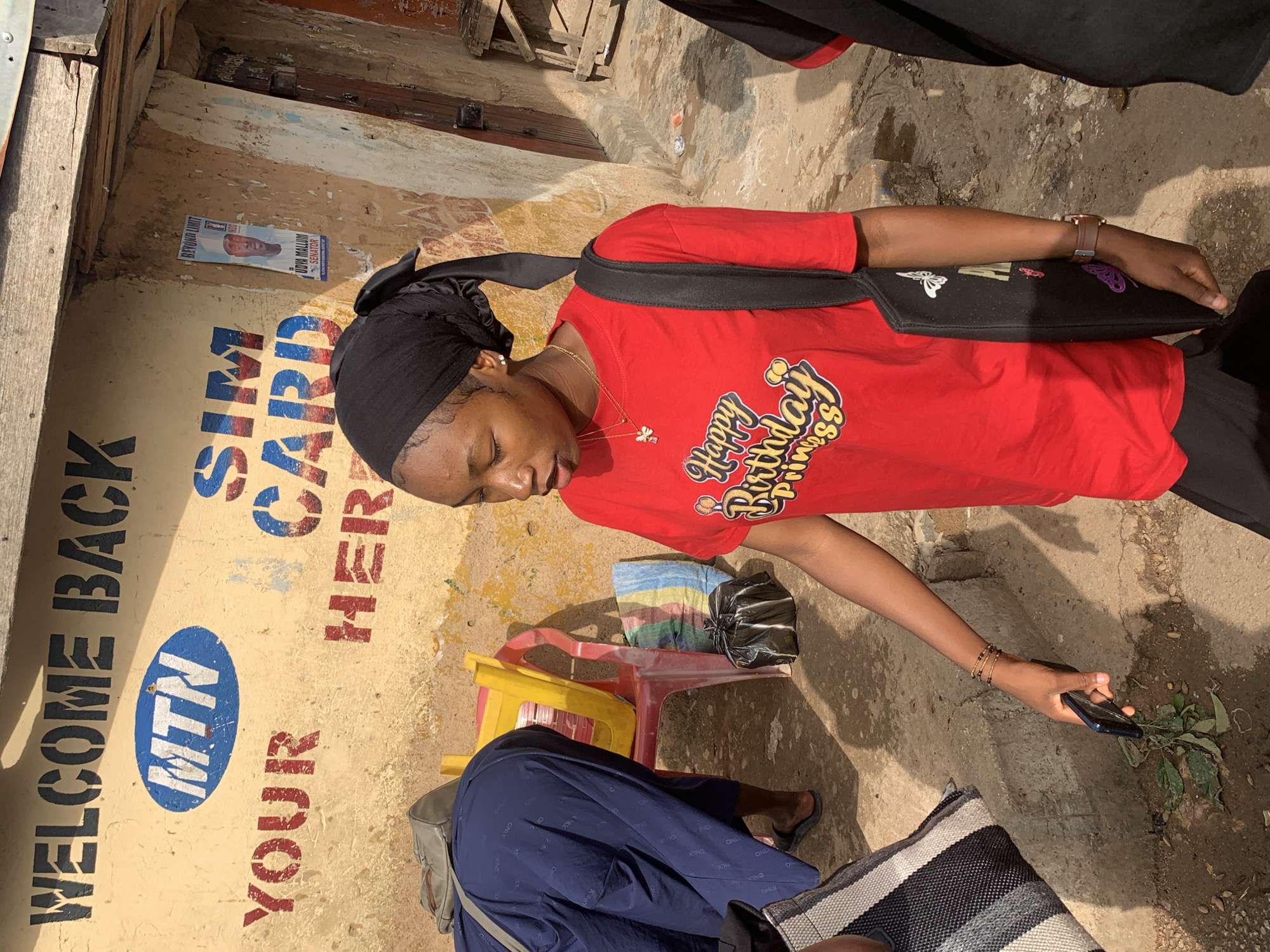 In Gura Top, Nigeria, a young woman wearing a black headscarf and a red t-shirt with "Happy Birthday Princess" printed on it is walking. She is holding a mobile phone in her right hand and carrying a black tote bag with butterfly designs over her left shoulder. Behind her, a person in a blue garment is bending over. The scene takes place outdoors in front of a building with yellow walls adorned with text and logos, including "WELCOME BACK", "MTN", "YOUR", "SIM CARD", and "HER". A poster featuring a man in a suit is also visible on the wall. In the background, plastic chairs and bags are scattered on the ground.