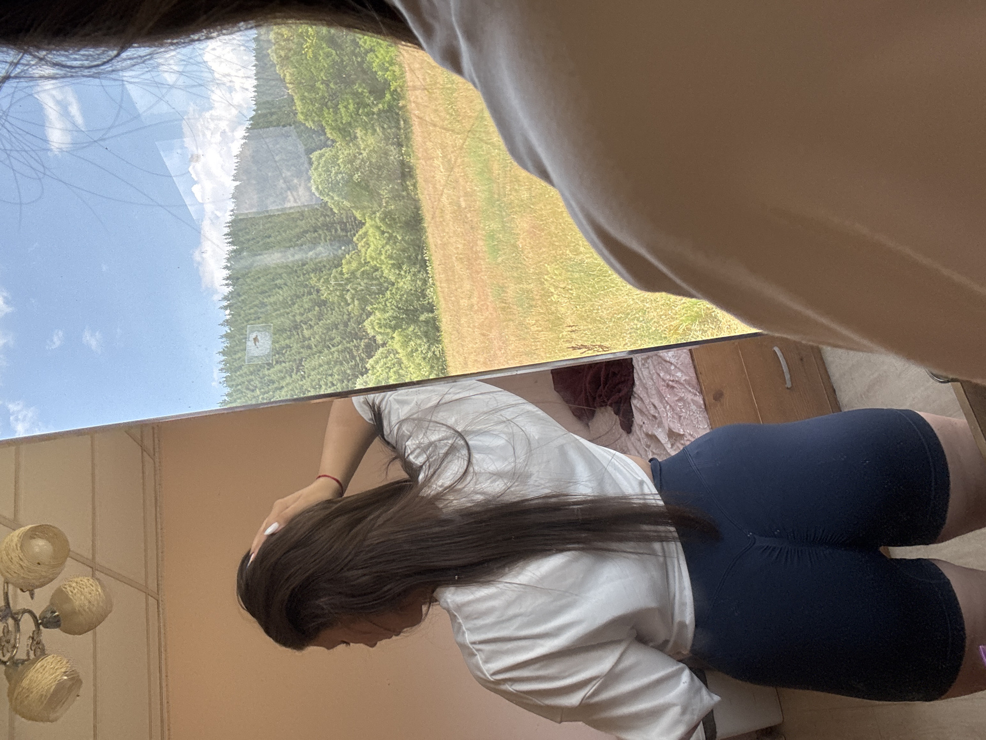 A woman is reflected in a mirror. She has long dark hair and is wearing a white t-shirt and dark blue shorts. Her left hand is touching her head. Behind her, a window reflects a landscape with a field, trees, and a sky with clouds. The room appears to be a bedroom, with a bed and a dresser visible in the reflection. The image appears to have been taken in Prodanovtsi, Bulgaria.
