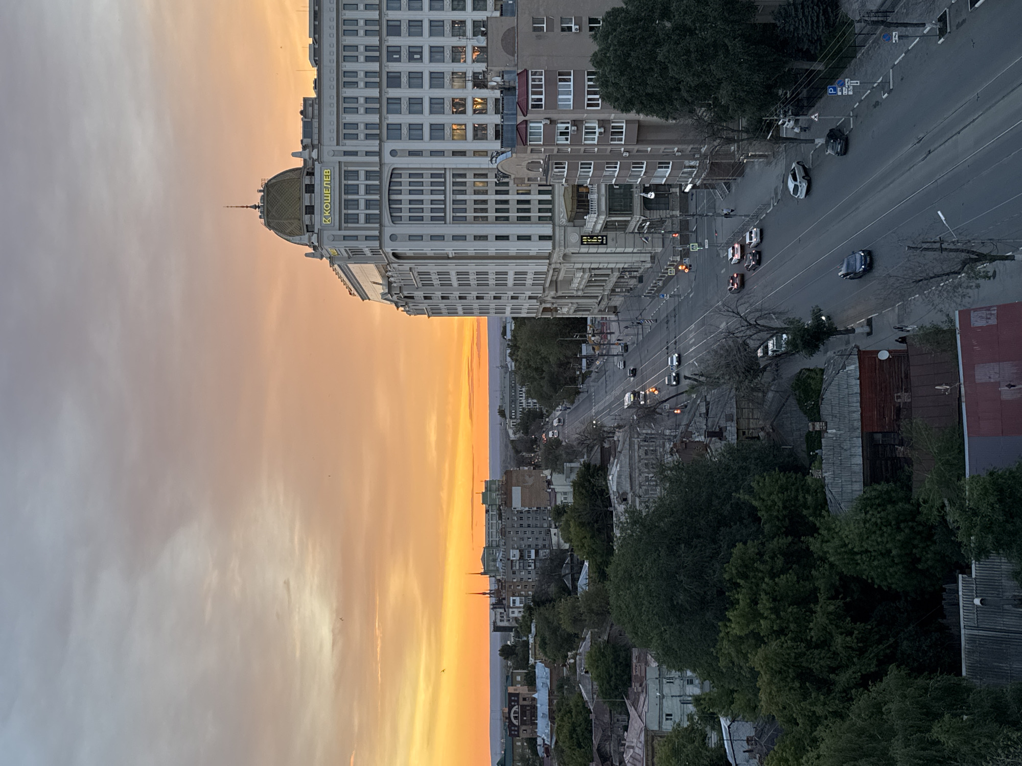 This is an aerial, wide-angle, eye-level shot of Novokuybyshevsk, Russia, at sunset. A wide, paved road with multiple lanes runs diagonally through the center of the frame, with traffic moving in both directions. Cars and a few pedestrians are visible on and near the road. On the right side of the road is a large, modern, white building with a golden dome and the Cyrillic lettering "К КОШЕЛЕВ" above its entrance. Adjacent to this is a multi-story residential building. On the left side of the road are older, lower-rise buildings and dense trees. In the distance, the sky is painted with vibrant orange and yellow hues of the sunset, with scattered clouds and a few birds flying. The overall scene depicts an urban environment transitioning from day to night under a colorful sky.
