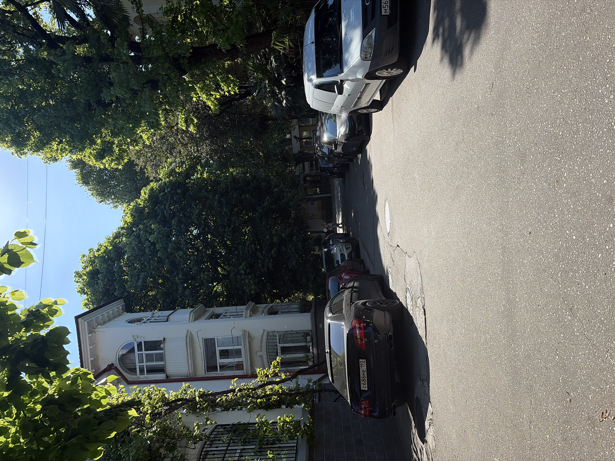 The image captures a sunlit street scene in Sochi, Russia, with the camera oriented vertically. On the right side of the frame, several vehicles are parked along the curb of an asphalt road. The most prominent is a white Mercedes-Benz Sprinter van, displaying a Russian license plate that reads M563TH. Behind it, a dark grey sedan is visible, bearing the license plate C686ME 193. The strong sunlight casts long, distinct shadows from the cars and overhead trees across the road, indicating either morning or late afternoon.
To the left, a multi-story white building with notable architectural features stands, characterized by its ornate details and a variety of windows, including distinctive arched and rounded ones. The street is heavily bordered by lush, dense green trees, some of which appear to be palm trees, hinting at a warm climate. A clear blue sky is visible in the upper left corner. The scene is devoid of people or explicit activity, conveying a sense of calm in a seemingly residential or mixed-use urban area.