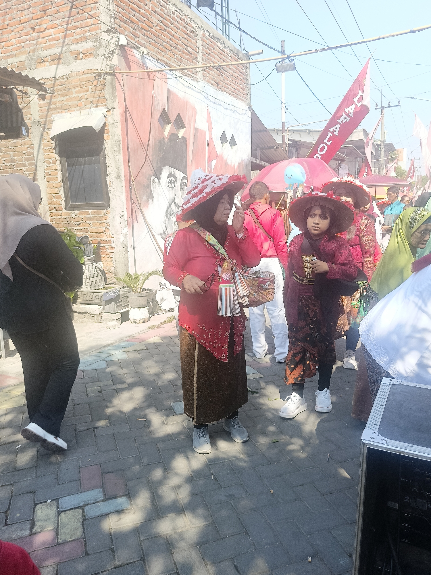 In Gedangan, Indonesia, on a sunlit day, a group of people are gathered outdoors on a paved walkway lined with brick buildings. A mural is visible on one of the buildings, depicting portraits. Several individuals are dressed in traditional Indonesian attire. Two women in the foreground wear large, ornate hats and vibrant, layered clothing, one in red and the other in dark red with gold accents. A younger person between them also wears a similar hat and a dark red, patterned outfit. A woman to the left is dressed in a hijab and black clothing, carrying a bag. In the background, other people are present, some holding flags. Red and white banners with text are visible, one with the word "DAHAT" prominently displayed. A pink umbrella with a blue toy hangs from it. The ground is paved with grey and colored interlocking bricks, with shadows from overhead structures cast across it. A black speaker box is positioned on the right side of the frame. The scene appears to be part of a cultural event or parade.