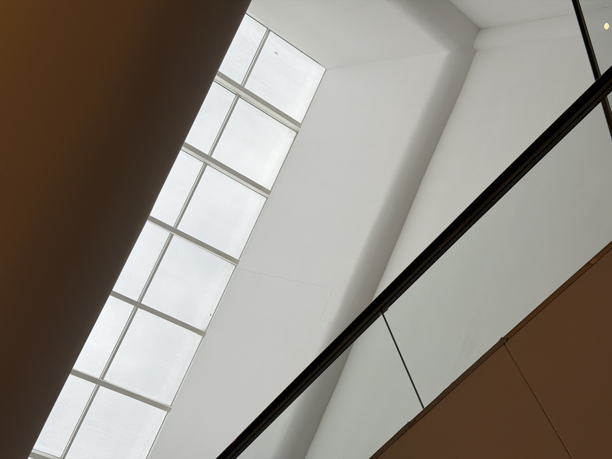 The image depicts an upward view of an interior space, likely a building in Moscow, Russia, given the architectural style. The scene features a large, angled window composed of multiple panes. Adjacent to the window is a white wall and an escalator with a glass barrier and a brown handrail. The architectural details suggest a modern design. No people are visible.