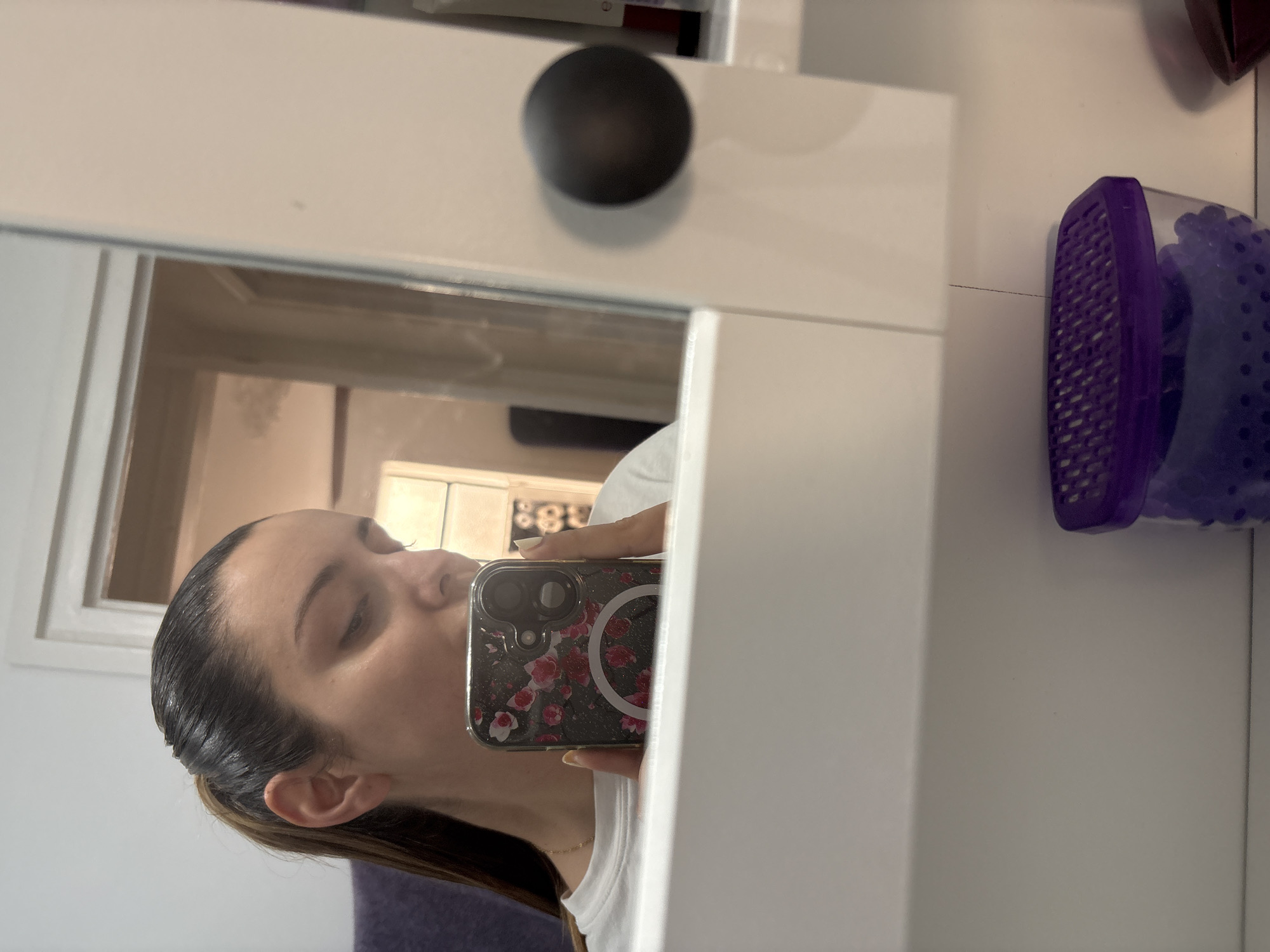 A woman is taking a selfie in a mirror. She is holding a phone with a floral-patterned case to her face. A purple object sits on a white surface to the right of the mirror. The mirror is attached to a white cabinet with a black knob. Through the mirror, a room is visible. The image offers no indication of the location in Barcelona, Spain.