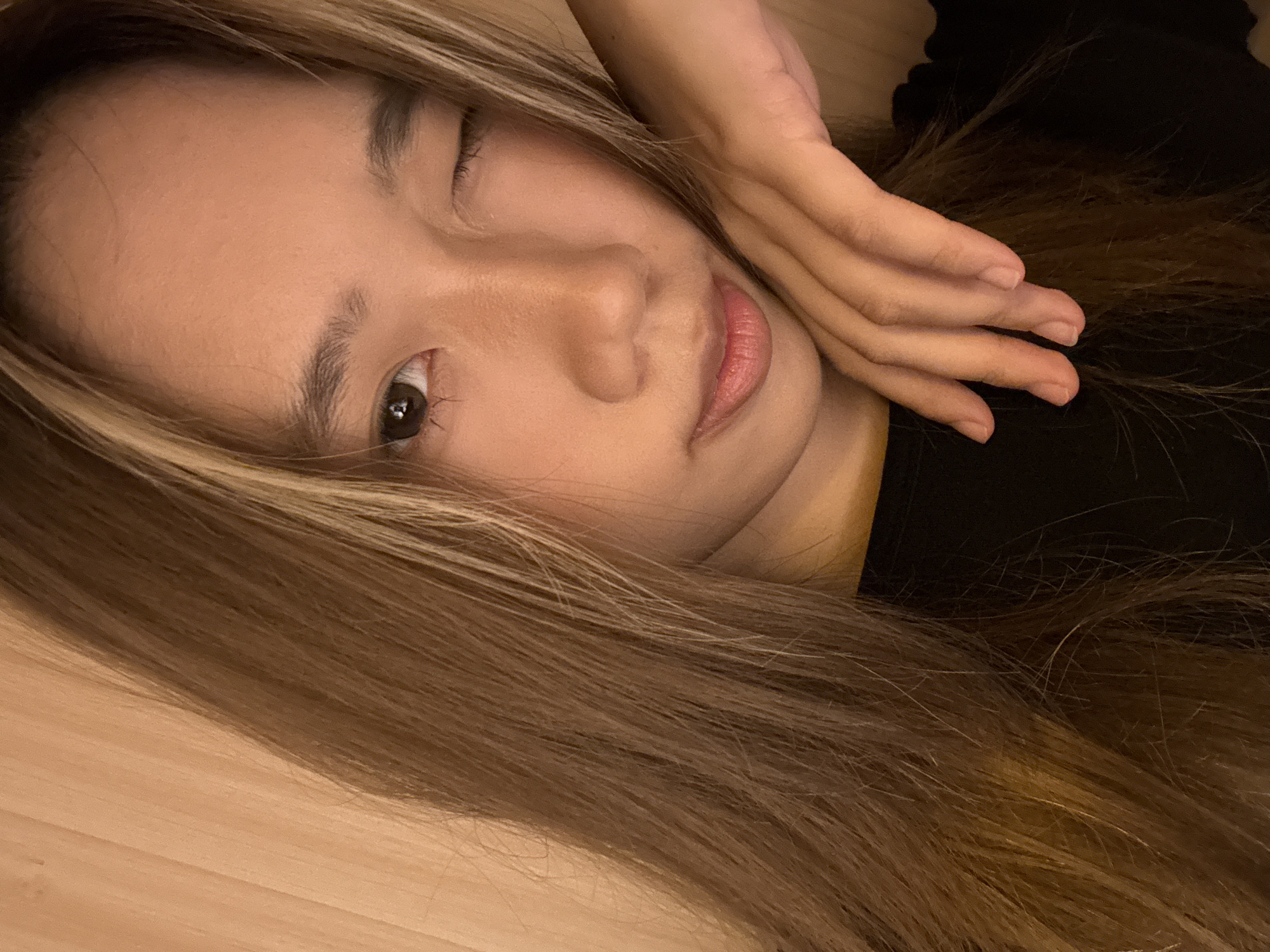 A close-up photograph shows an Asian woman with brown hair with blonde highlights lying down. She is turned to her left, with one eye open and the other closed in a wink or partial squint. Her right hand is resting on her face. She appears to be wearing a black top. The background is blurry, with some visible woodgrain texture suggesting a floor or other wooden surface. No location in Yuzhong District, China, is evident.