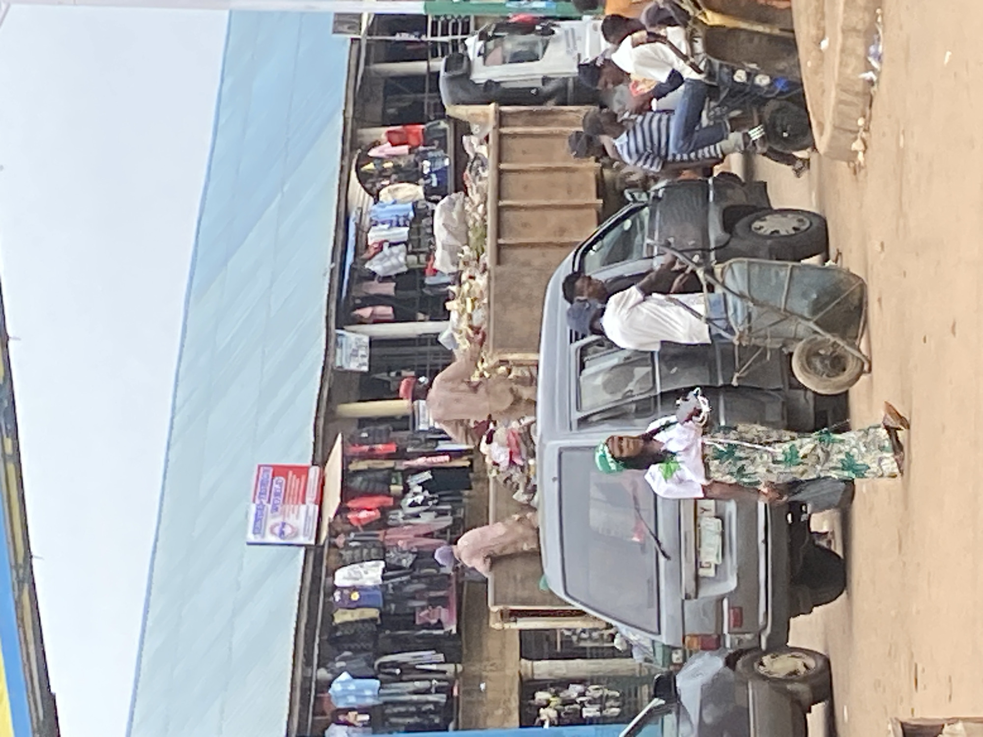 In Bukuru, Nigeria, a busy street scene unfolds near a market. A large, loaded truck with its bed full of debris sits prominently in the center, suggesting a clean-up or collection operation. In front of the truck, a grey minivan is parked. A woman, dressed in a white shirt and a patterned green and white skirt, walks across the foreground, carrying a small item in her hand. Behind her, several men are gathered, some appearing to be pushing or attending to a trolley with wheels. Further back, on the left side of the image, a stall displays a variety of clothing items hanging on racks, indicating a commercial area. The overall atmosphere appears to be one of everyday commerce and activity in a bustling urban environment.