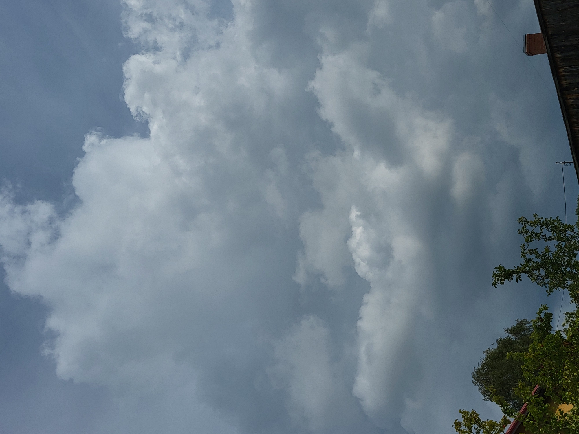 The image shows a dark, cloudy sky with some lighter clouds dispersed throughout. The lower part of the image features the tops of trees and a wooden structure, possibly a roof, with a brick chimney. The scene appears to be outdoors, in a rural or suburban setting. The lighting suggests it might be overcast or late afternoon.