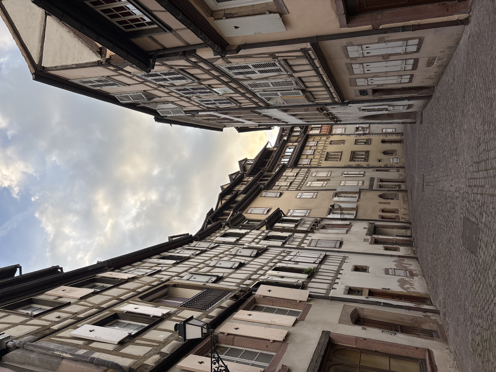 This is a low-angle, wide shot of a narrow cobblestone street in Colmar, France, lined with traditional half-timbered buildings. The architecture features exposed wooden beams against lighter colored stucco walls, with many windows covered by white wooden shutters. The sky above is partly cloudy, with the soft light suggesting either early morning or late afternoon. The street appears empty of people, giving the scene a quiet and tranquil atmosphere. The perspective draws the eye down the street, which curves slightly out of view. The overall impression is one of historical charm and picturesque European urban scenery.