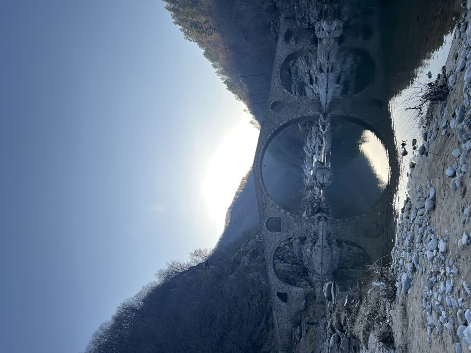 The image displays a multi-arch stone bridge spanning a calm river in a mountainous valley near the city of Diadovtsi, Bulgaria. The bridge, constructed from dark masonry, features a prominent central arch flanked by two smaller arches and additional minor arches extending towards the left bank. A strong, low-lying light source, likely the sun, is positioned behind the bridge, casting a bright glare and reflecting significantly on the river's surface.
Steep, tree-covered hillsides frame the valley. The vegetation on these slopes appears largely bare or sparse, indicative of a non-summer season. The hillside on the left is predominantly in shadow, while the right hillside exhibits patches of direct illumination from the light source.
The river beneath the bridge is placid, mirroring the bridge structure and the bright sky. The riverbed in shallower areas is visibly rocky, with numerous exposed boulders. The immediate foreground consists of a rocky and sandy riverbank with scattered patches of dry vegetation. One individual, dressed in dark clothing, is visible standing on the left riverbank near the water's edge. The sky above is clear and light blue, transitioning to a brighter tone near the light source.