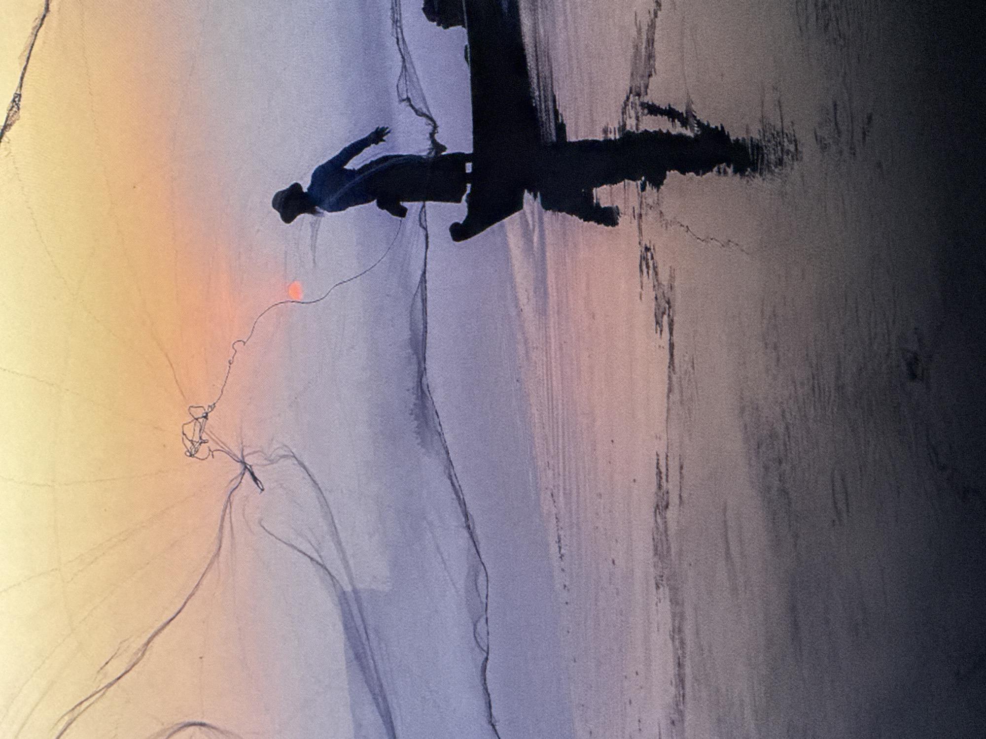 The image depicts a silhouette of a lone fisherman casting a net from a small boat. The fisherman, wearing a hat, is in the process of throwing a large net, which is spread out in a wide arc against a twilight sky. The sky transitions from a warm orange and yellow at the top to a soft purple and pink near the horizon. The water surface reflects the colors of the sky and the dark silhouette of the boat and fisherman. The overall scene evokes a sense of tranquility and the quiet pursuit of a livelihood. The location is not identifiable as Saratov, Russia, nor is there any indication of a city in the image.