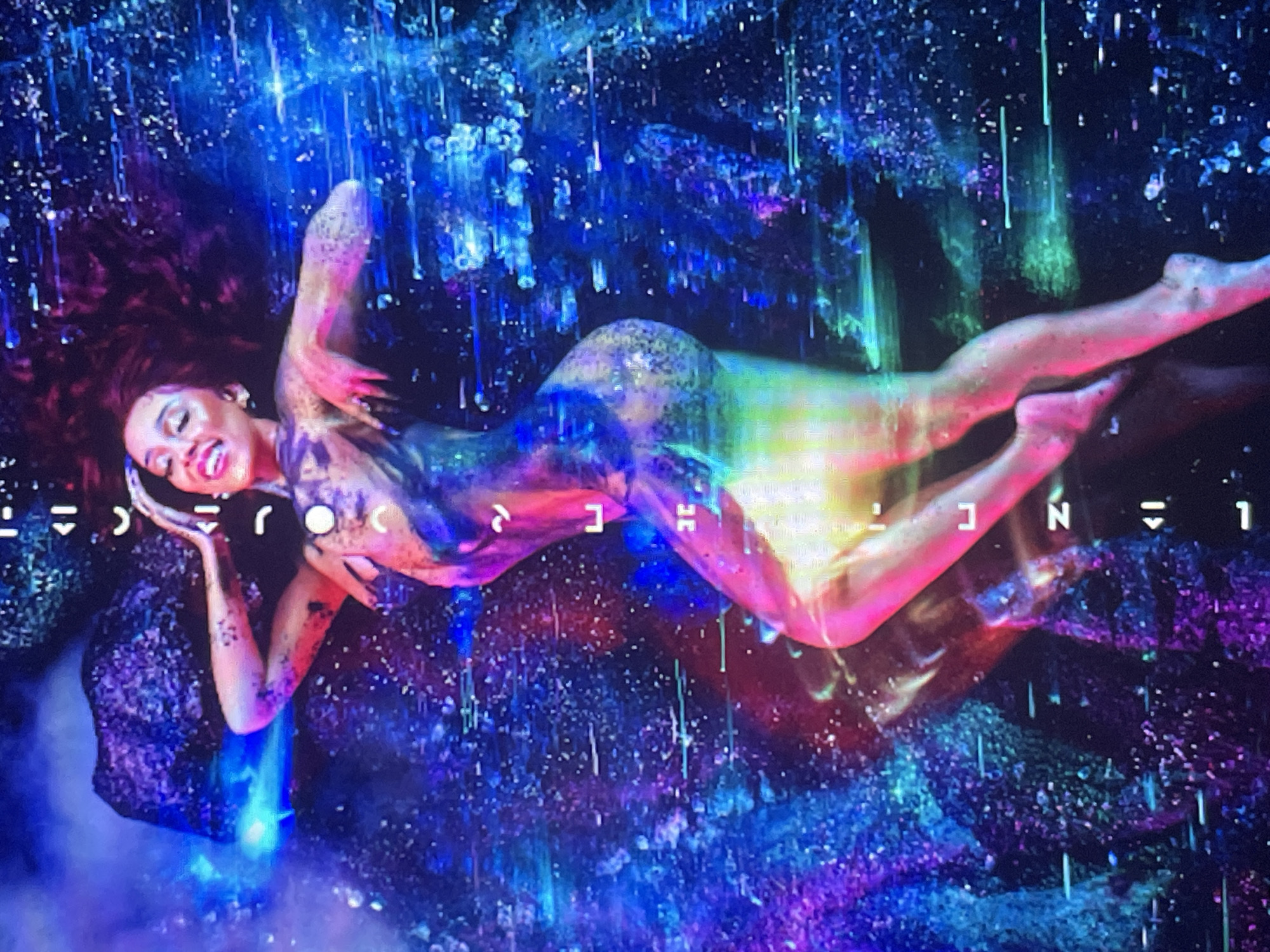 The image depicts a woman, possibly Doja Cat, floating horizontally in a vibrant, abstract, cosmic or deep-sea environment. Her head is positioned to the left, and she looks towards the viewer with a joyful, serene smile. Her dark hair flows around her head, and her body is adorned with iridescent, multi-colored paint or fabric in shades of purple, blue, green, pink, and yellow. Her limbs are gracefully extended, suggesting a state of weightlessness.
The background is a dynamic tapestry of deep blues, purples, and greens, filled with countless sparkling points of light and vertical luminous streaks, evoking a sense of stars, nebulae, or bioluminescent underwater phenomena. These elements create an ethereal and surreal atmosphere around the central figure.
Scattered throughout the background are stylized, abstract symbols or glyphs that appear to be part of the artistic design. These symbols, resembling characters such as '(', 'J', 'O', 'C', '<', 'N', '=', and '1', contribute to the otherworldly aesthetic.
The scene is highly stylized and artificial, offering no discernible time of day or weather conditions. The woman's pose and expression convey a sense of wonder and contentment. There are no location cues within the image itself that suggest Sofia, Bulgaria.