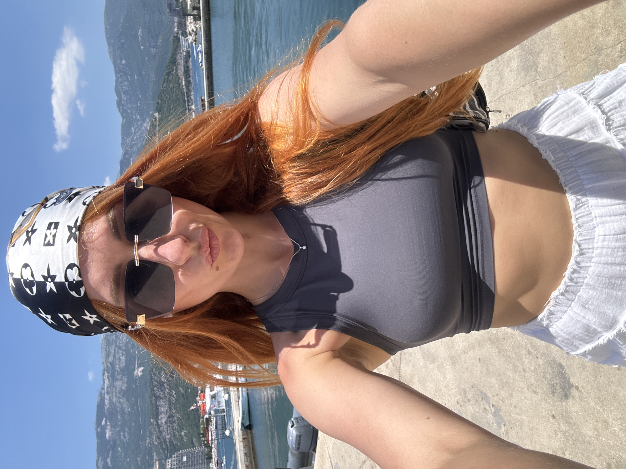 A woman with long, red hair, wearing a grey crop top, white ruffled shorts, a patterned bandana, and large sunglasses, takes a selfie outdoors. She is standing on a concrete surface next to a body of water, with boats and mountains in the background. The location appears to be a harbor in Budva, Montenegro, as indicated by the distinct architecture of the buildings on the hillside and the characteristic mountainous terrain. The sky is clear and blue with some light clouds.