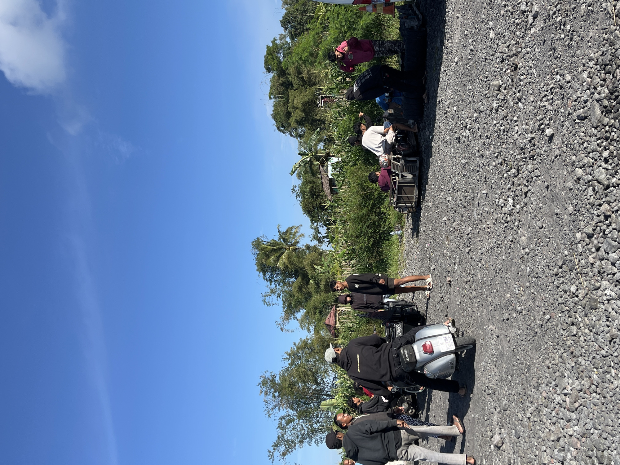 The image depicts a group of young men gathered on a dirt road in Oro-oro Ombo, Indonesia, with a lush, green landscape and a clear blue sky in the background. Several individuals are seen socializing around motorcycles, with some seated on a modified motorcycle sidecar. The road surface is covered in gravel, and there is dense vegetation, including trees and crops, bordering the area. The overall atmosphere suggests a casual outdoor gathering.