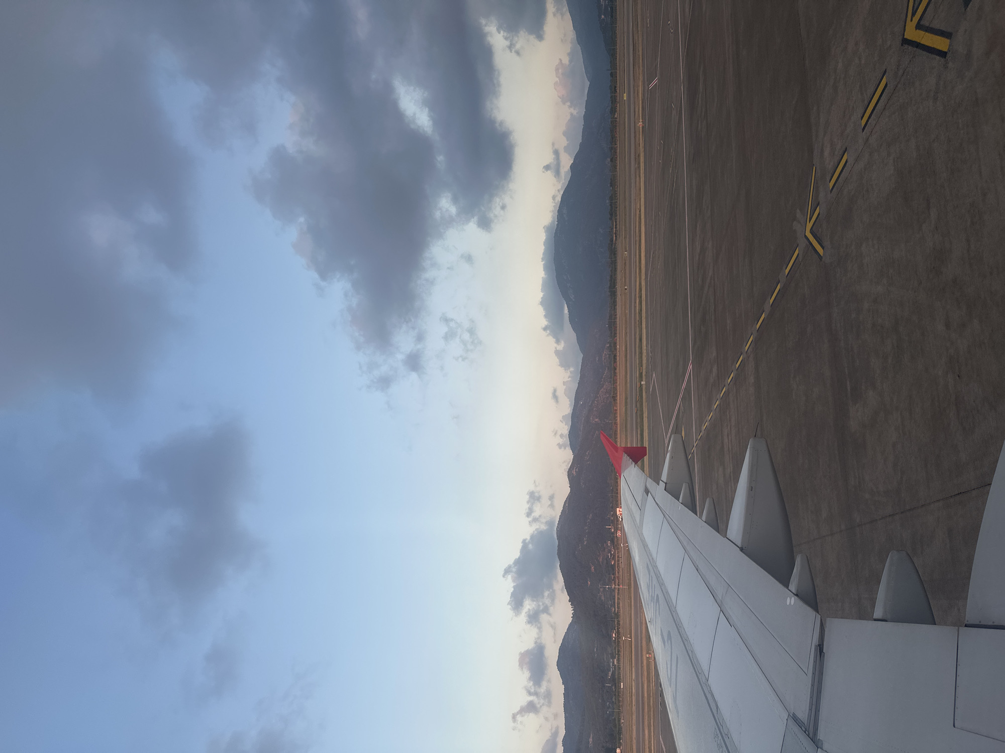 This image presents a detailed view from an aircraft window at Dalaman Airport (DLM) in Dalaman, Turkey, captured during either dawn or dusk. The left side of the frame features the light gray wing of an airplane, equipped with a prominent red wingtip (sharklet) on its outer edge. Faint lettering or a logo is discernible on the upper surface of the wing closer to the fuselage.
The foreground and midground to the right of the aircraft wing display dark gray asphalt or concrete airport ground, marked with distinct yellow dashed lines forming taxiway indicators and directional arrows. Additional white lines are visible further back on the airfield. Portions of the ground in the distance exhibit a warm, golden illumination.
Across the horizon, a range of dark, rugged mountains dominates the background, appearing sparsely vegetated. A faint line of human settlement or infrastructure, including subtle lights, is visible at the base of these mountains between the airfield and the peaks.
The sky transitions from a light blue with warm, soft hues near the mountain horizon to a darker, muted blue-gray in the upper portion. Scattered gray clouds, some with lighter edges catching distant light, are distributed throughout the upper half of the sky. No individuals are explicitly visible within the scene.