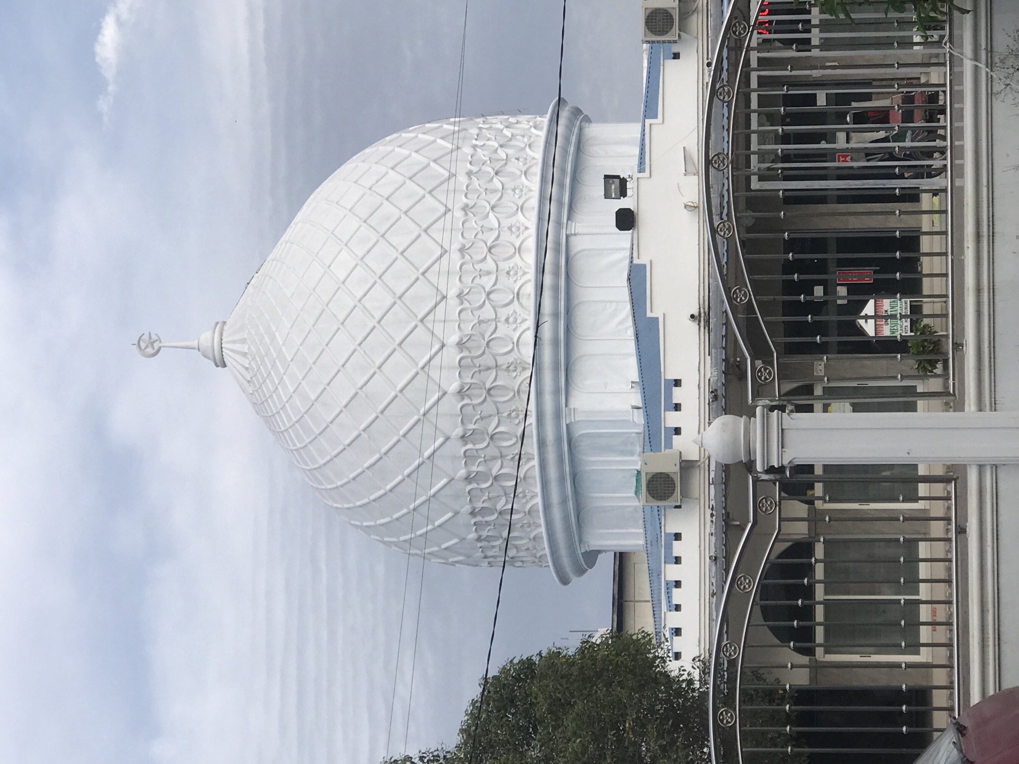 The image depicts the Baiturrahman Mosque, located in Lhokseumawe, Indonesia. The mosque features a large, white dome with a crescent moon and star finial at its apex. The dome is decorated with a geometric pattern. The building also has a white facade with blue accents and is visible behind a metal fence. An air conditioning unit is mounted on the exterior wall. The sky is overcast with light gray clouds. Power lines are visible in the foreground. Green foliage is also visible near the bottom of the frame.