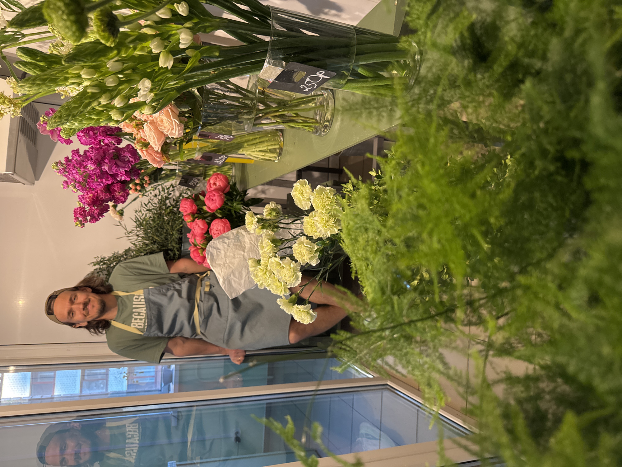 A man wearing a green shirt and a gray apron with the word "BECAUSE" is standing in what appears to be a flower shop. He is positioned behind a counter displaying various flowers in vases. The flowers include white lilies, pink roses, purple stocks, and pink peonies. There are price tags on some of the vases. The scene is partially obstructed by green ferns. The image is taken indoors near a window, and the man's reflection can be seen in the window. The location is Izhevsk, Russia.