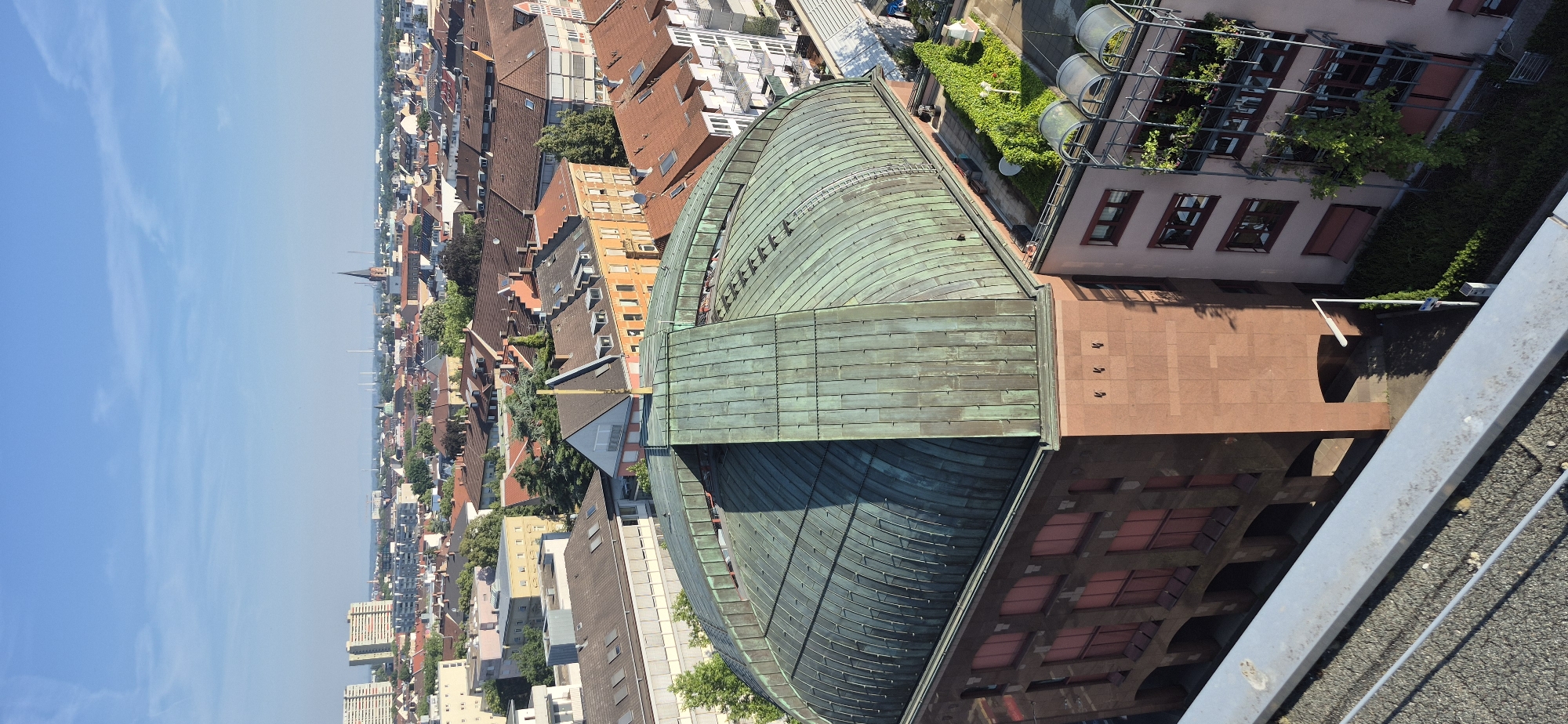 This is a high-angle, full-shot photograph capturing a cityscape under a clear blue sky, likely taken from a tall building. The foreground is dominated by a weathered, grey metallic railing and a rough, asphalt-like surface. In the midground, a large building with a distinctive curved, copper-green roof is prominent. The building has a reddish-brown facade with arched openings at its base and several rectangular windows with reddish-pink frames on its upper levels. To the right of this building, another structure with a lighter facade and multiple windows is visible, featuring balconies with greenery. The background reveals a dense urban sprawl of buildings with red-tiled roofs, interspersed with trees. In the far distance, taller, modern buildings and industrial structures, including a church spire, are discernible, suggesting the urban center of Karlsruhe, Germany. No people are visible in the frame.