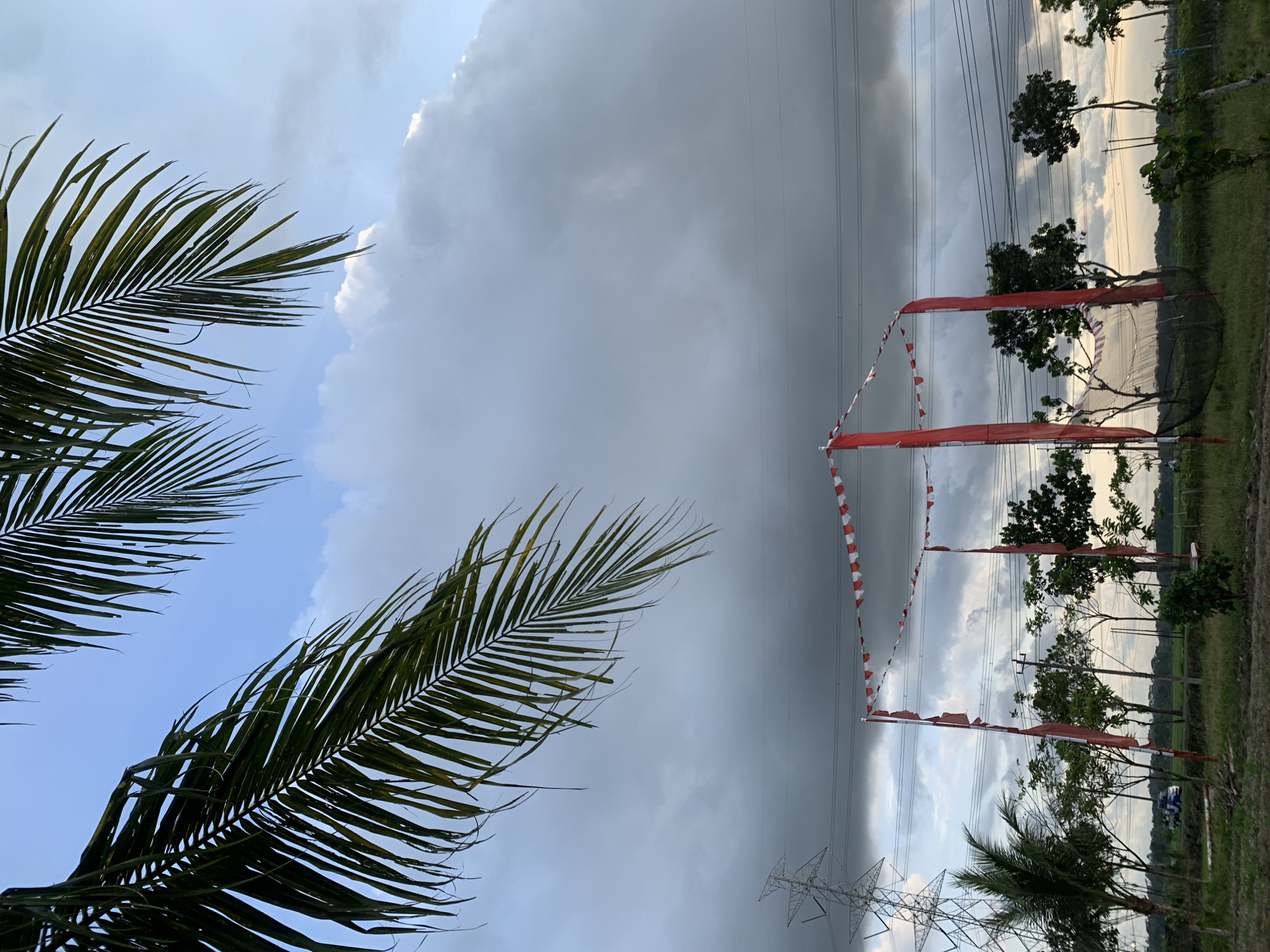The image depicts a scene in Karangkemiri, Indonesia. Dominating the foreground are palm fronds framing the sky. The sky itself is a mix of blue patches and imposing, dark storm clouds. An electricity pylon is visible, along with power lines stretching across the scene. Red and white bunting or flags are strung up, likely for a local celebration, near some small trees. The background features a field or area of low vegetation with some larger trees visible in the distance.