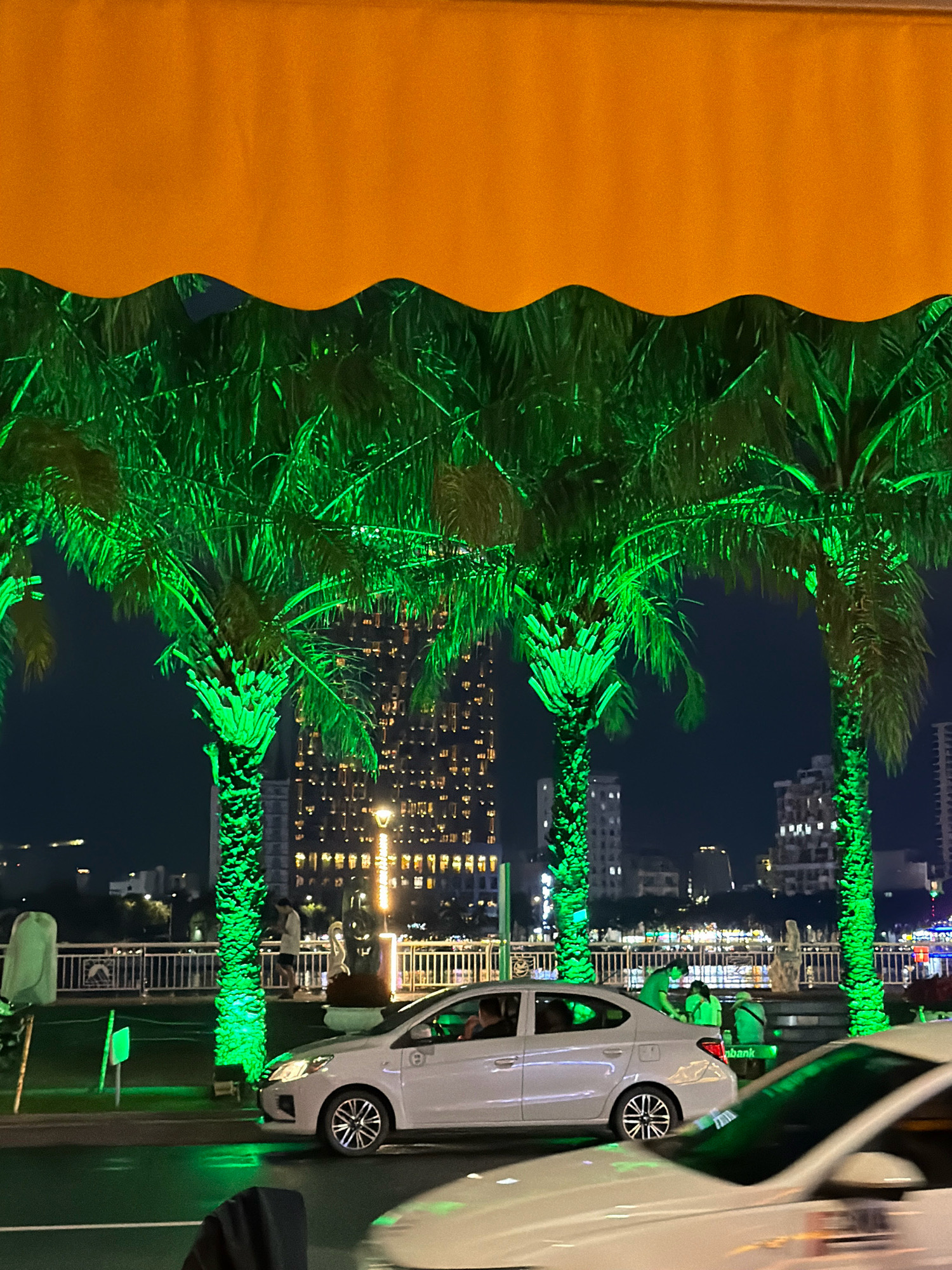 In this nighttime photograph, palm trees are illuminated with vibrant green lights, creating an atmospheric scene. In the foreground, a white car is visible, its headlights casting a glow on the wet street, suggesting recent rain. Behind the palm trees, a cityscape is illuminated, with tall buildings featuring numerous lit windows. A pedestrian bridge is also discernible, with railings and streetlights. The overall setting appears to be a waterfront area in Đà Nẵng, Vietnam, possibly along the Han River, given the presence of illuminated buildings and the general ambiance of a bustling urban environment at night. The bright orange awning at the top of the image frames the scene, adding a contrasting color element.