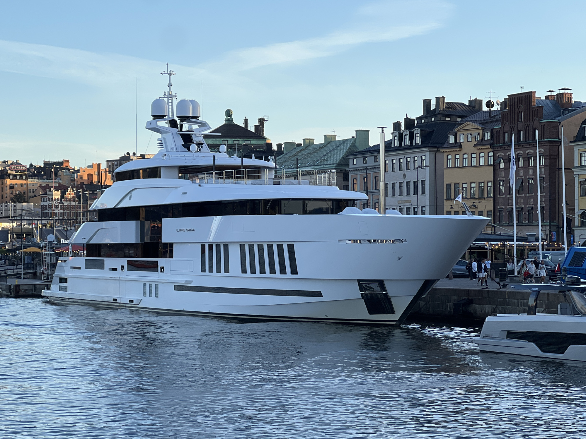 A large, white yacht named "LIFE SAGA" is docked in the water, in front of a row of old, multi-story buildings with distinctive architecture. The buildings appear to be in the Gamla Stan district of Stockholm, Sweden, with their red-tiled roofs and facades visible. The sky is a clear blue with a few wispy clouds. Several people are visible on the dock behind the yacht, some appearing to be walking or talking. Another smaller, modern boat is partially visible in the foreground on the right. The water is calm with gentle ripples. The overall scene suggests a picturesque harbor setting in a historic European city.