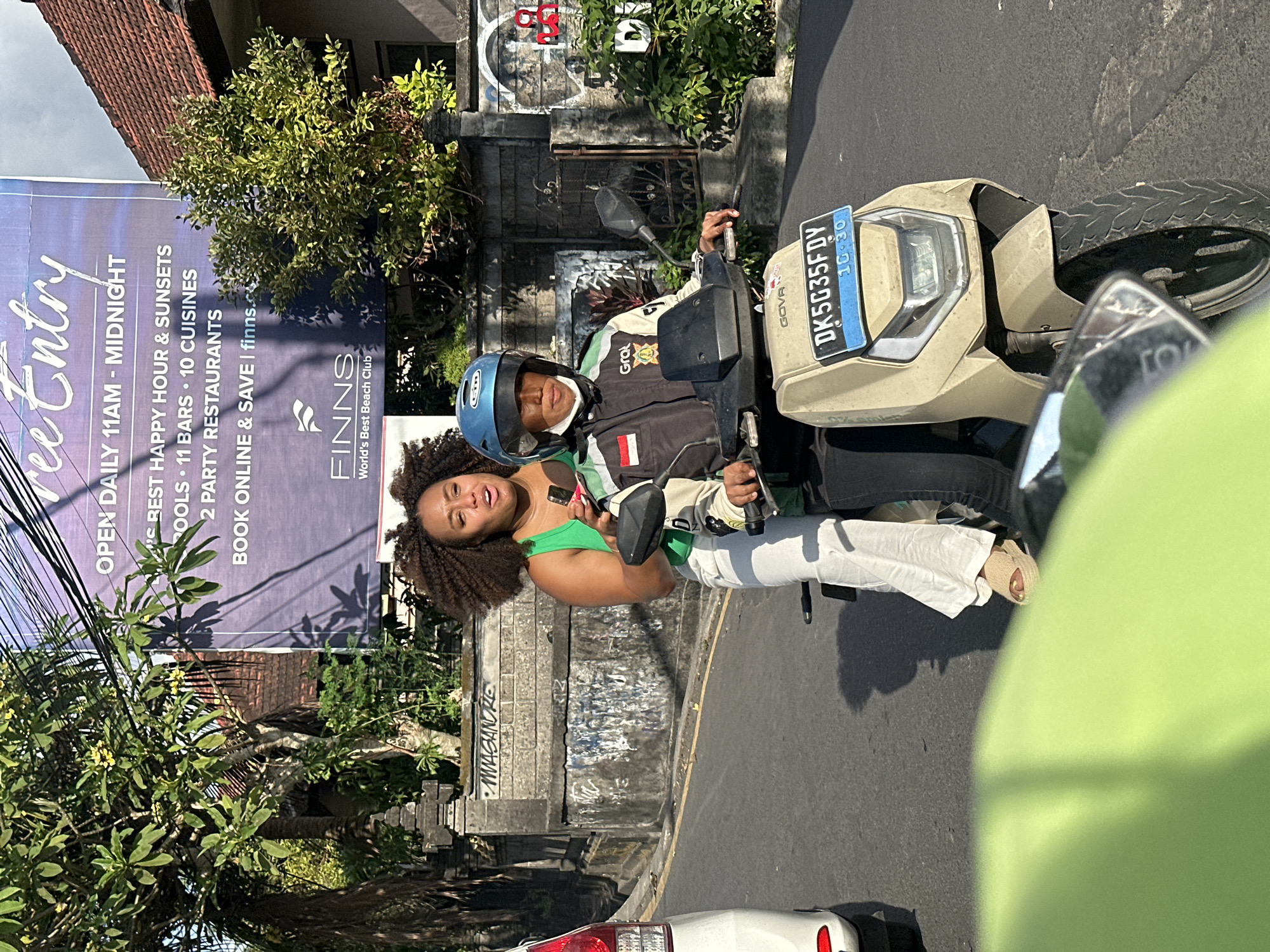 The image captures a street scene in North Kuta, Indonesia, where two individuals are riding a scooter. The person in the front, wearing a blue helmet and a black and green jacket with "Grob" visible, is operating the scooter. The person in the back, a woman with voluminous curly hair wearing a green tank top and white pants, is holding a mobile phone in her right hand. They are traveling on a paved road under a clear sky, with buildings and lush green foliage visible in the background. A large blue banner with white text, advertising "Finns World's Best Beach Club," is prominently displayed on a building. The scooter has a license plate that reads "DK5035FDY" and displays "10-30". The overall atmosphere suggests a typical day in a tropical urban environment.