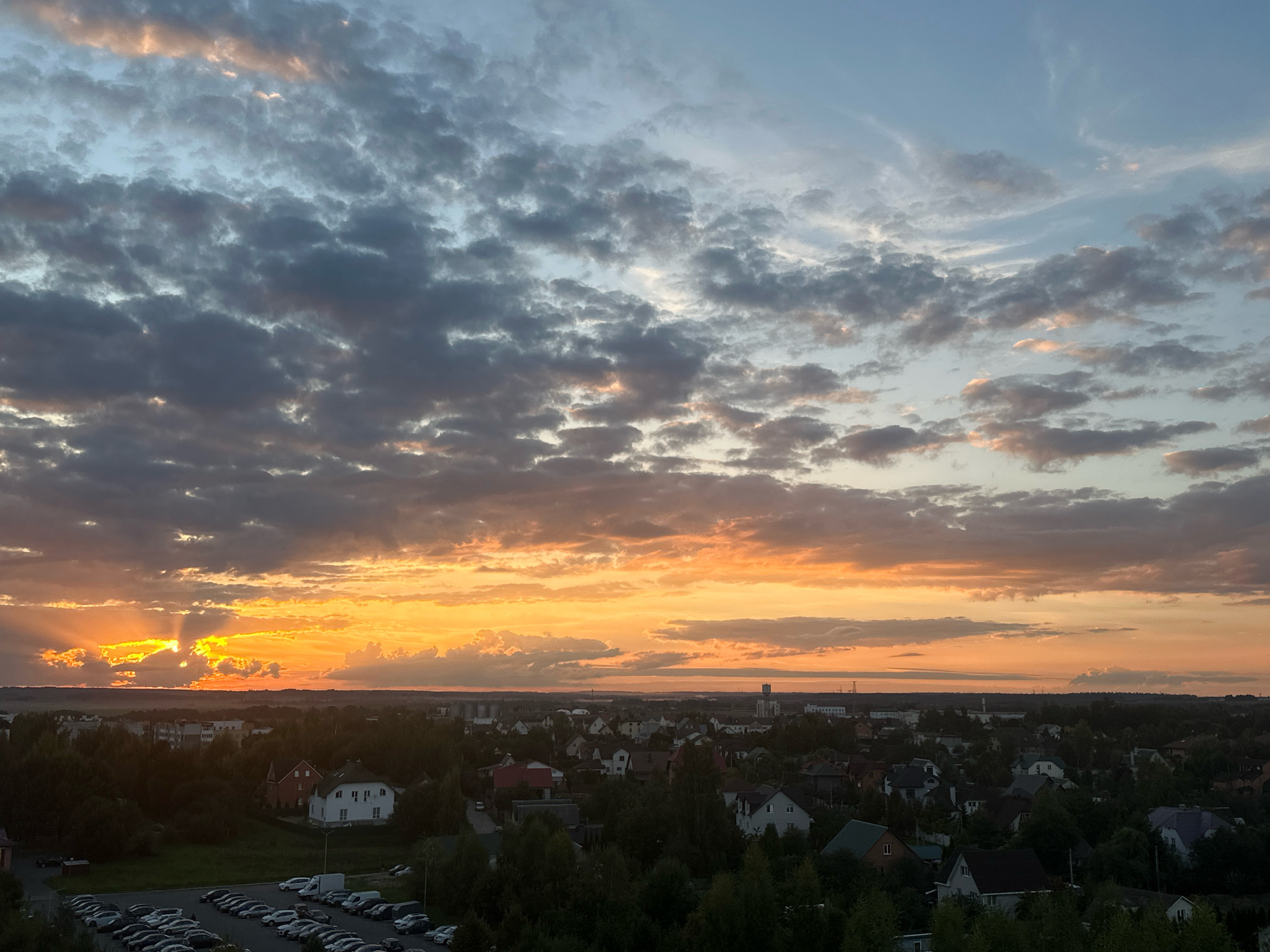 This is an aerial, wide-angle, landscape photograph taken at dusk or dawn over the Shchomyslitsky Sel'sky Soviet, Belarus. The sky is filled with dramatic, textured clouds. The sun is low on the horizon, casting a brilliant orange glow through the clouds on the left side of the frame, with rays of light fanning out across the scene. The foreground consists of a dense, dark green forest and a suburban residential area with numerous houses. Some houses are visible with red and white facades, and others with darker roofs. A parking lot filled with numerous cars, including a white van, is situated in the lower left corner of the image. A few taller buildings, possibly industrial or residential complexes, are visible in the distance, along with a tower-like structure. The overall atmosphere is serene, with the dramatic sky contrasting with the quiet, shadowed landscape below.