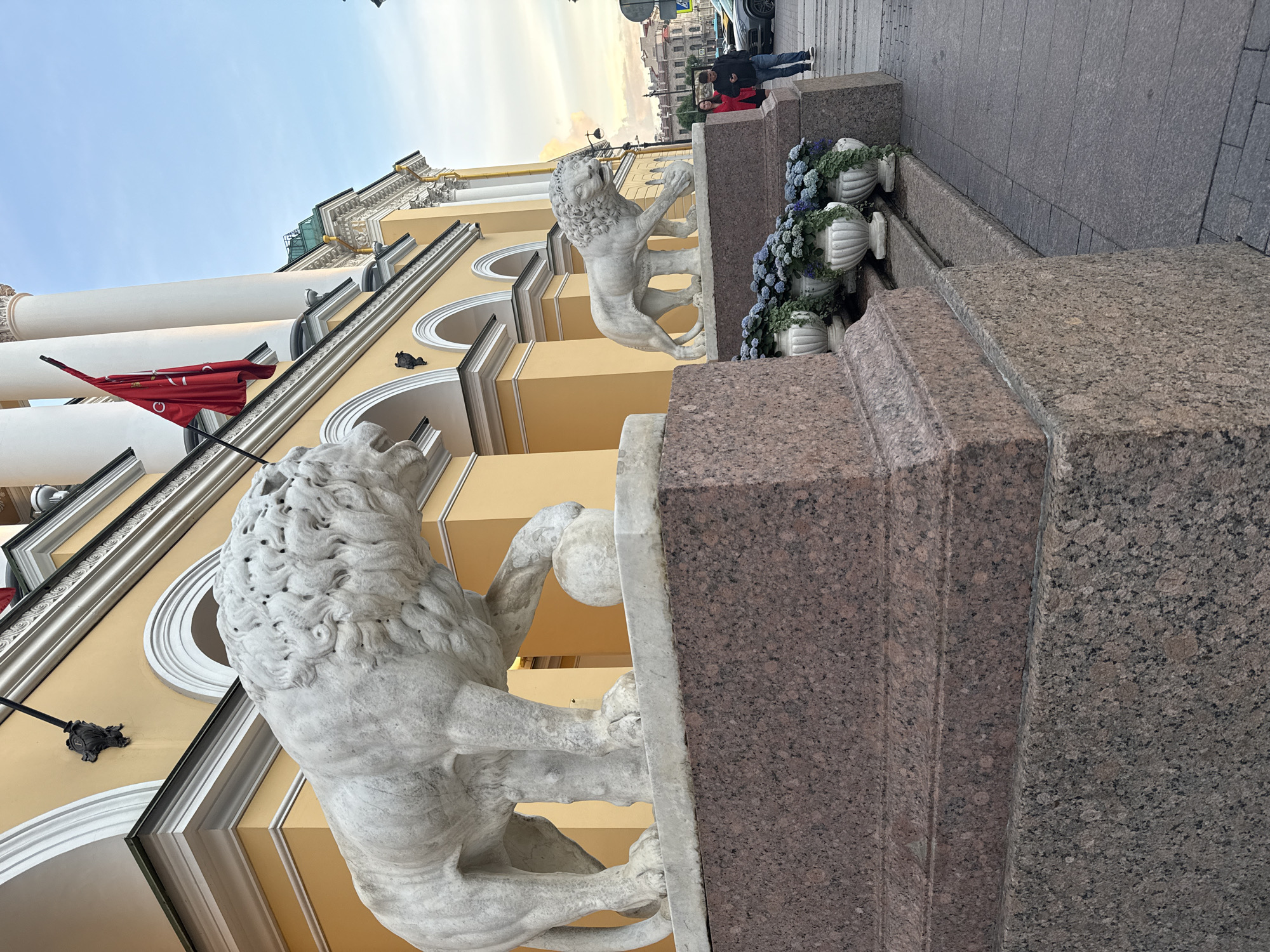 The image depicts the Lion statues at the Lion Bridge in Saint Petersburg, Russia. The statue in the foreground, larger and closer to the viewer, is a white marble lion with its paw resting on a ball. It stands on a granite pedestal with a reddish-brown hue. Behind it, another, smaller lion statue is visible, also white marble with a ball under its paw. The statues are positioned in front of a yellow building with classical architectural elements, including arched entrances and white columns. A red flag with a crescent symbol is visible flying from the building. In the background, a sidewalk is visible, with two individuals standing to the right of the smaller lion statue. One person, wearing a red jacket, is looking towards the camera, while the other, wearing a dark jacket and blue jeans, is looking at their phone. Further in the background, there are city buildings and a street with vehicles, suggesting an urban environment. Several decorative planters with blue flowers and green foliage are placed along the steps leading up to the statues. The sky above is a pale blue with some clouds, indicating it is likely late afternoon or early evening.
