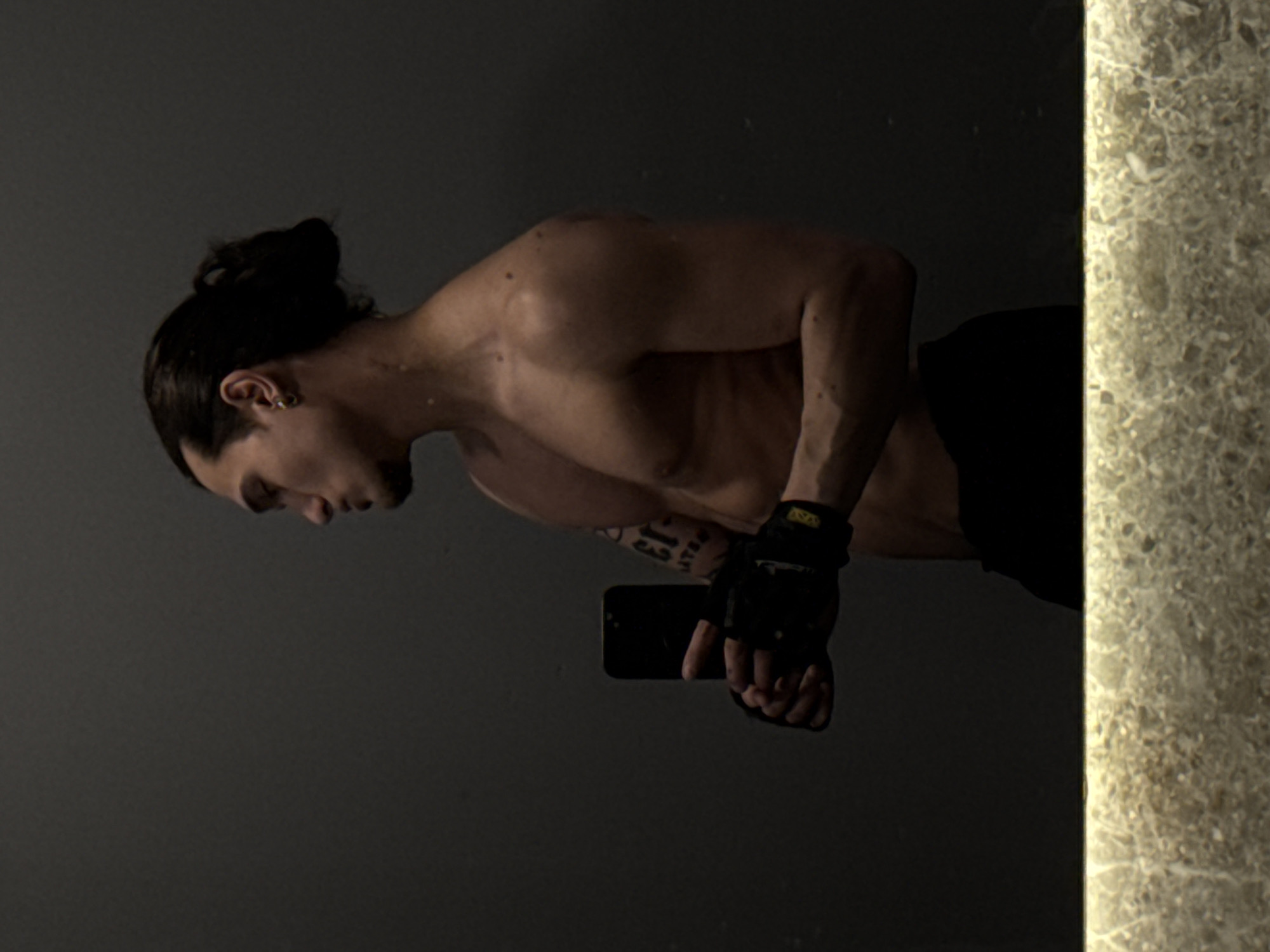 A medium shot captures a muscular man taking a mirror selfie indoors. He is shirtless, revealing a toned physique, and his dark hair is tied back in a bun. His head is tilted downwards and slightly to his left, suggesting a focused or pensive expression. He wears a black fingerless glove on his left hand, with faint yellow lettering, possibly "VNA," visible on the strap. A black smartphone is held in this gloved hand, pointed towards the mirror, indicating the self-portrait. Tattoos are visible on his left forearm, but the text is indistinguishable.
The background primarily consists of a dark, reflective surface, likely a large mirror. To the right of the frame, a textured, light-colored surface, resembling a stone or marble pillar, provides a visual contrast to the dark backdrop. The lighting is subdued, creating shadows that accentuate the man's musculature. The setting appears to be an indoor space, possibly a gym or a private room. There are no overt cues for the time of day or weather, and the man's demeanor conveys a sense of concentration or seriousness.