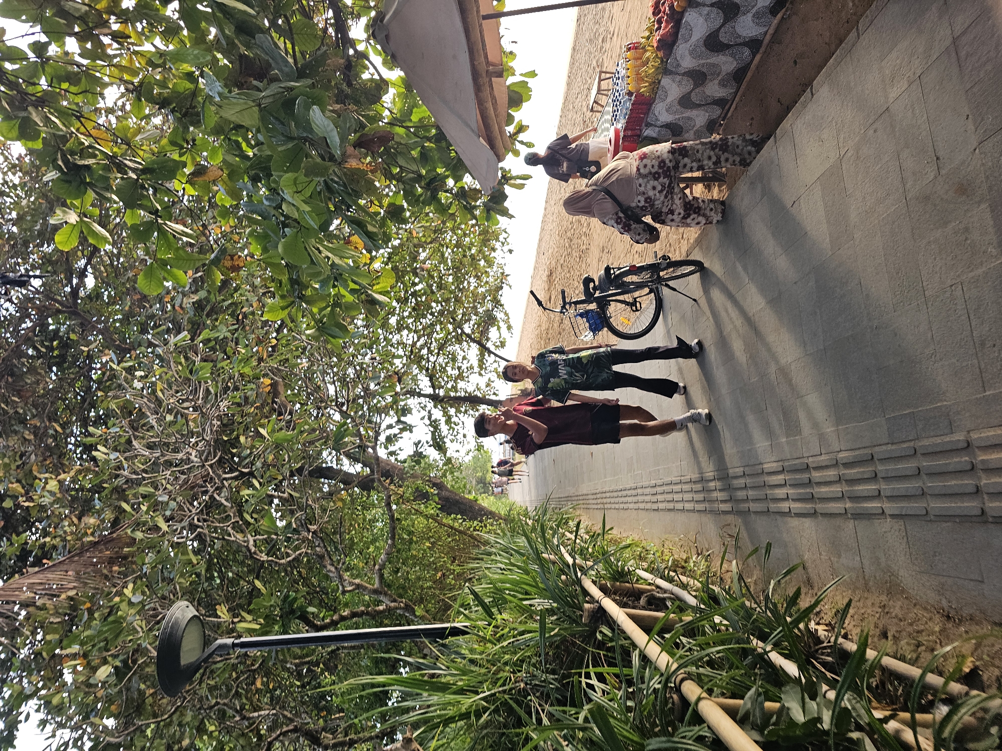 The image shows a promenade by a sandy beach in Denpasar, Indonesia. Two young people, a male and a female, are walking on the promenade. The male is wearing a maroon t-shirt and black shorts, while the female is wearing a green t-shirt and black trousers. A bicycle is parked on the promenade next to them. Further down the promenade, a person wearing a headscarf and patterned trousers is seated next to a stall selling fruits and drinks under a large umbrella. Another person is standing behind the stall. The promenade is lined with lush green vegetation on one side and the beach on the other. Trees provide shade over the promenade. The scene appears to be during the daytime, possibly late afternoon, given the angle of the sunlight and the shadows.