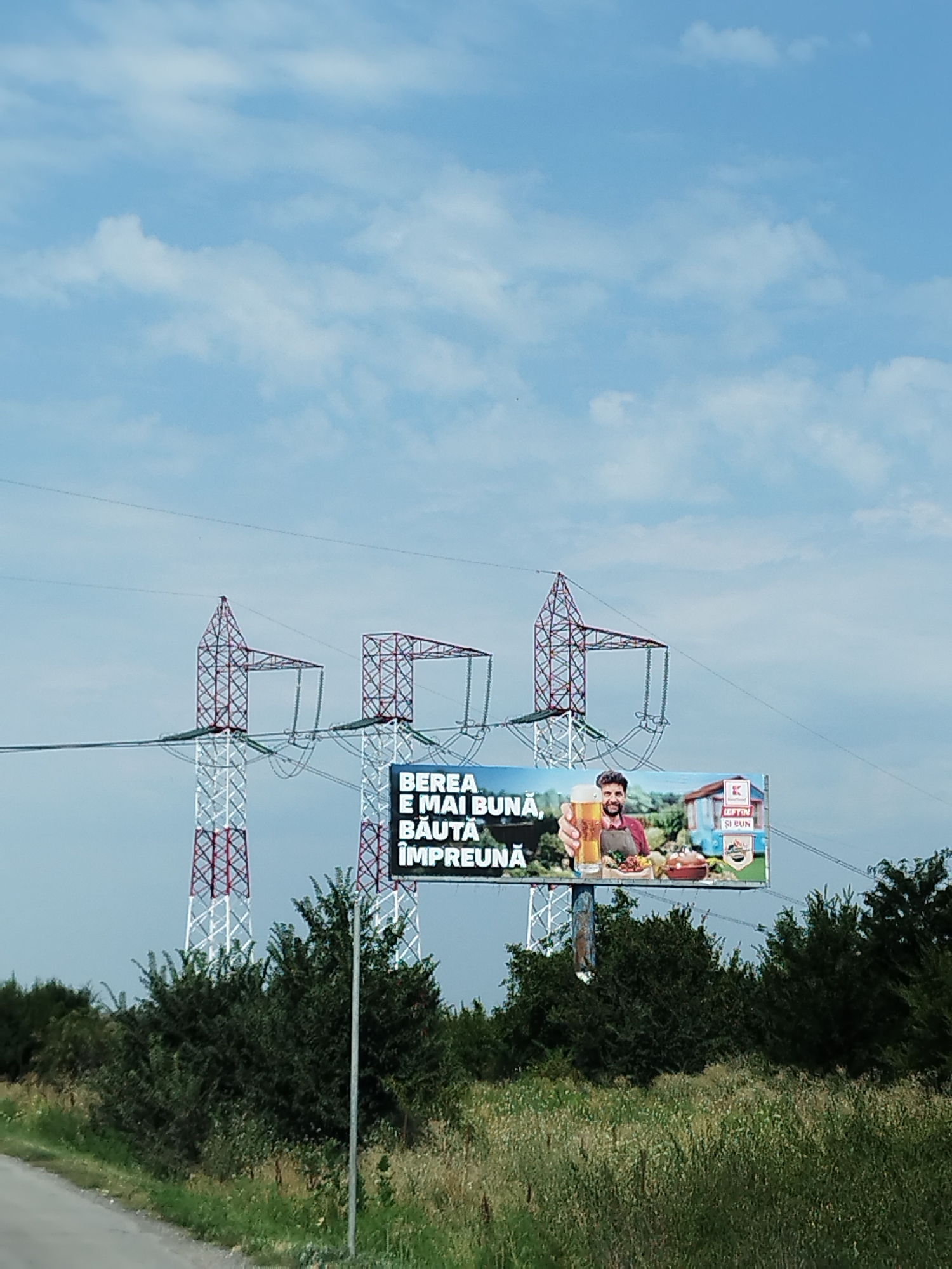 This image captures a billboard advertising beer, situated in the city of Joița, Romania. The billboard features the text "BEREA E MAI BUNĂ BĂUTĂ ÎMPREUNĂ," which translates to "Beer is better drunk together." A man is depicted holding a large glass of beer, alongside a spread of food and a grill, suggesting a social gathering. To the right of the main advertisement, there is a smaller inset showing a house with the text "BFTIN SI BUN," possibly indicating a product or brand name. The billboard is supported by large, red and white lattice-work electricity pylons. In the foreground, lush green trees and dry grass line a paved road, with the sky a clear blue with scattered clouds. The overall scene is a roadside advertisement promoting shared enjoyment of beer in a natural setting.