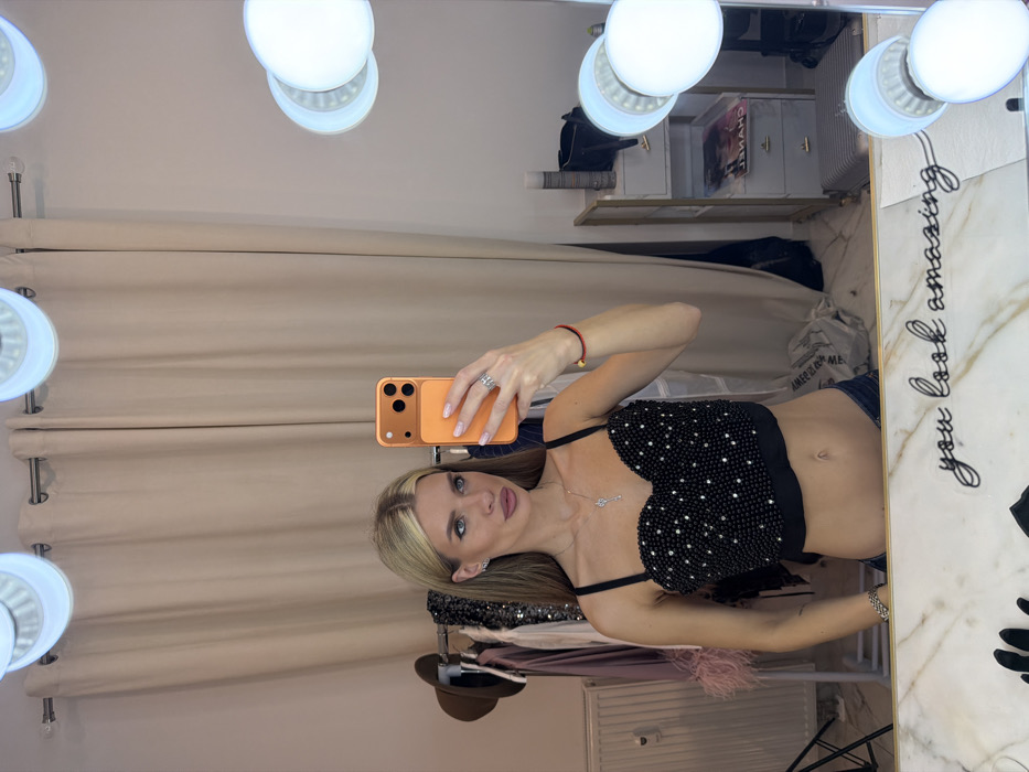 A woman poses in a mirror selfie, showcasing a black, sequined crop top. She holds an orange smartphone, revealing manicured nails and rings. Her blonde hair is styled with side part, and she wears subtle makeup. The mirror is surrounded by vanity lights. The reflection shows a clothing rack, shelving with items, and a printed message stating "you look amazing."