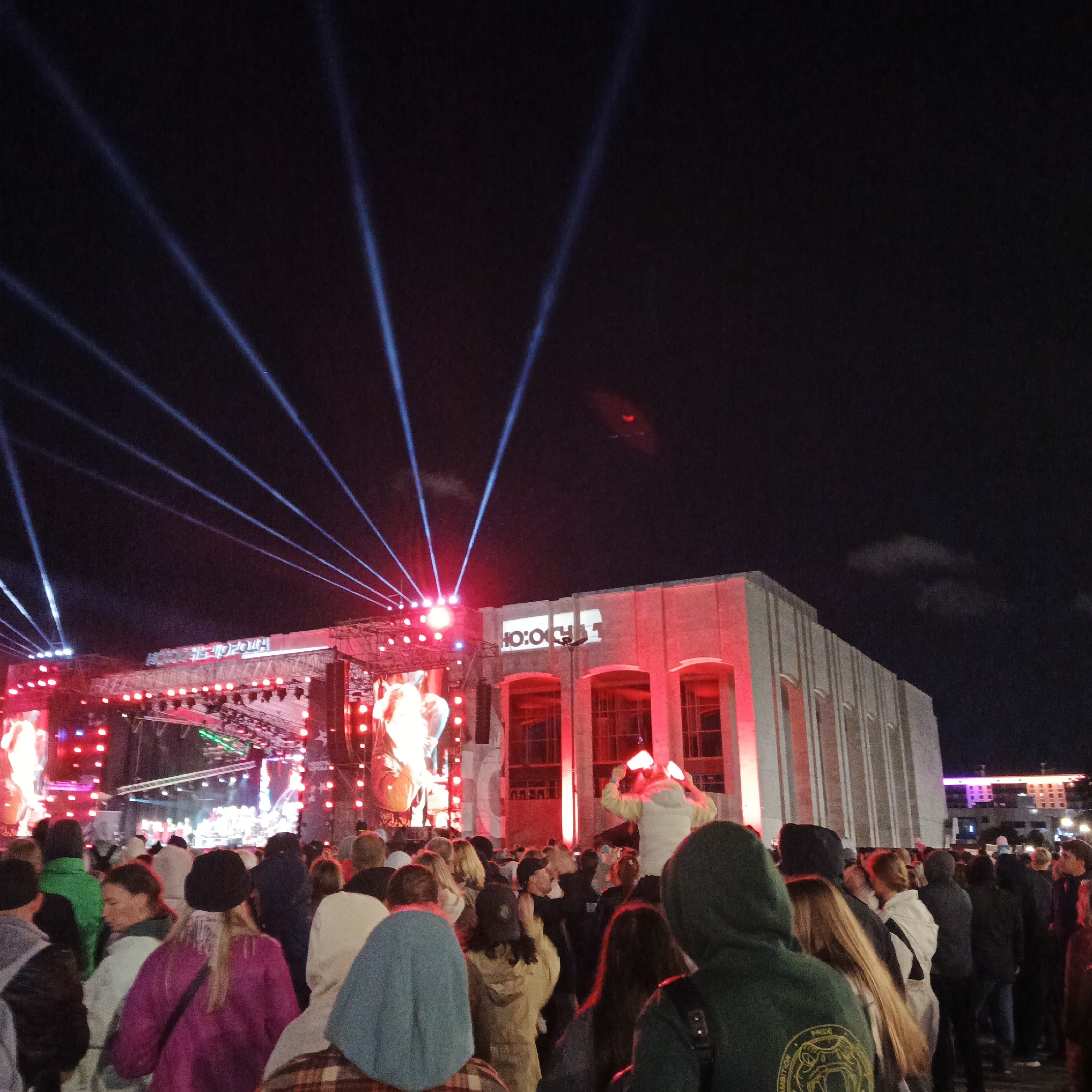 An outdoor concert is taking place at night in the city of Perm, Russia. A large crowd of people are gathered, facing a brightly lit stage with several performers visible. Searchlights are projected into the dark sky, creating dramatic beams of light. A building with illuminated windows and red architectural lighting stands to the right of the stage. The overall atmosphere appears festive and energetic.