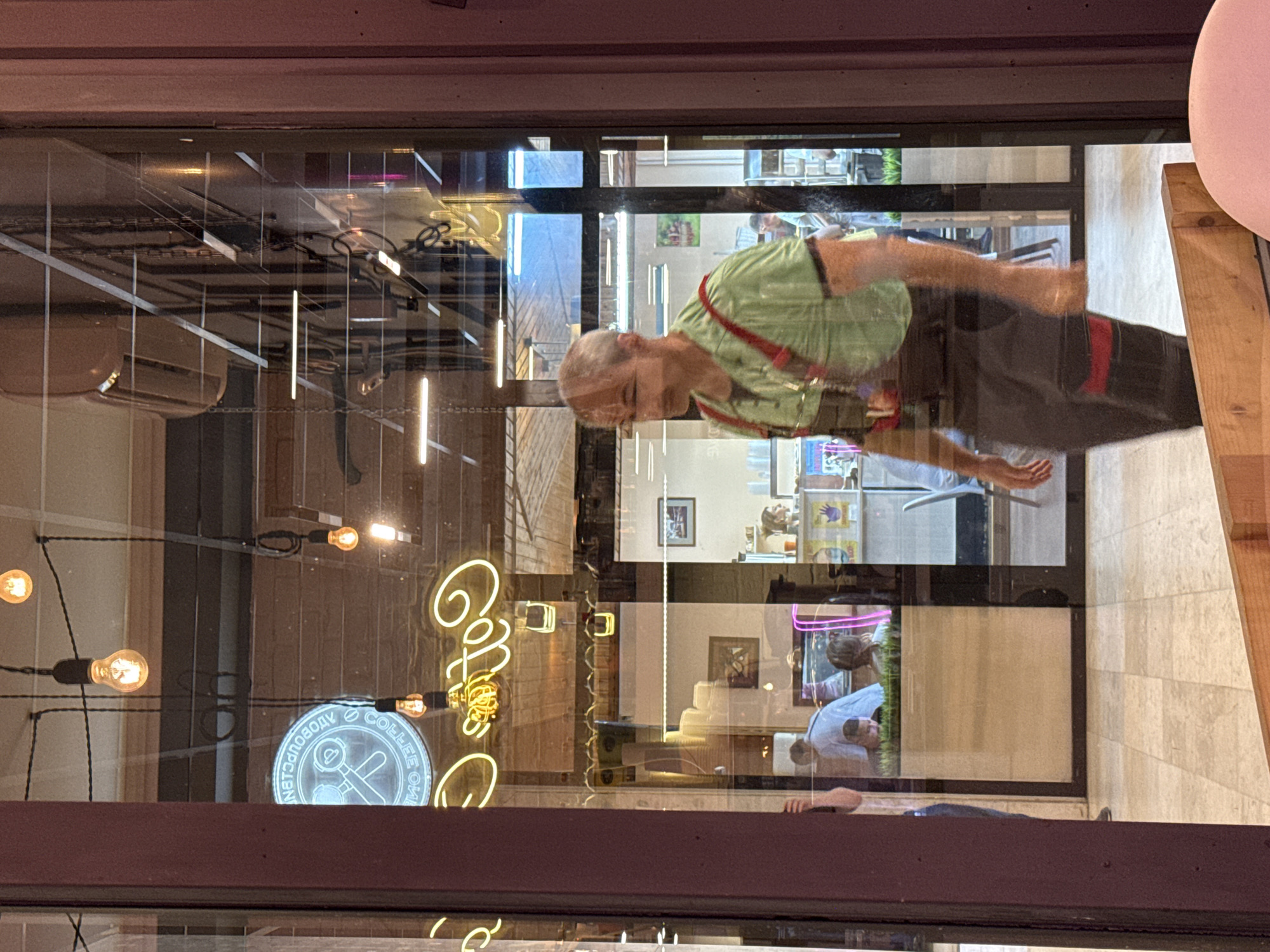 The image depicts a man in a green shirt and dark overalls walking through a doorway, reflected in a window. The man is gray-haired and appears to be looking to his left. Behind him, inside what appears to be a cafe or shop, other people are visible. A neon sign with the word "Coffee" in yellow letters and a circular logo with a coffee-related image in blue are illuminated in the window, suggesting the establishment. The scene is set in Moscow, Russia. The floor is tiled, and a wooden plank is visible in the foreground. The lighting suggests it might be daytime or early evening.