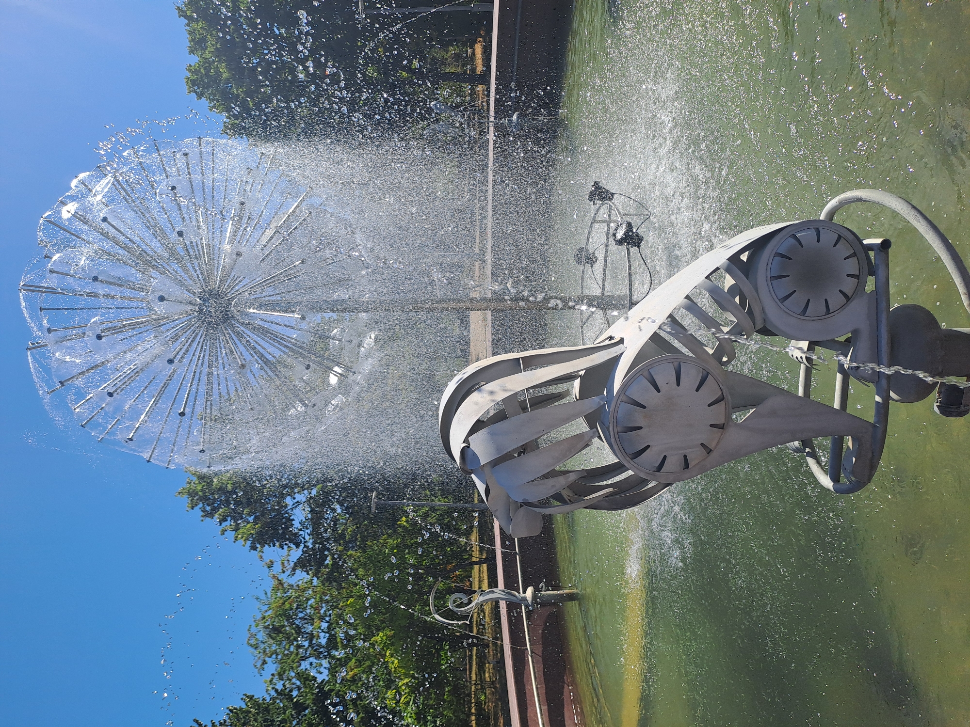 The image shows a dandelion-shaped fountain in a park in Chișinău, Moldova. The fountain is made of metal and has water spraying from it in a circular pattern. In the foreground, there is a metal sculpture of an abstract bird. The background features green trees and a clear blue sky. The water in the fountain is a murky green color, and the sun is shining brightly, creating reflections on the water's surface.