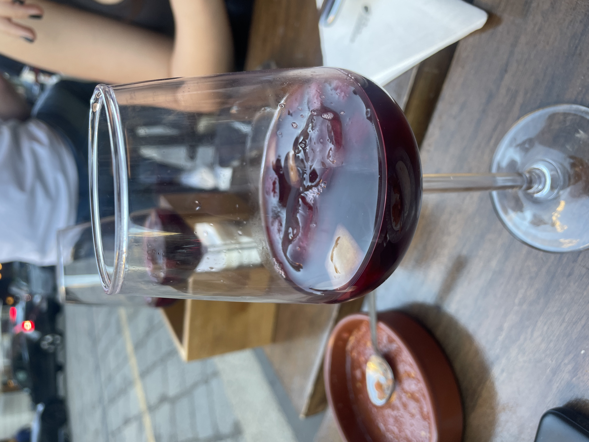 A glass of red wine with floating fruit is the primary focus of the image. The glass is positioned on a wooden table. Nearby, there is a small brown dish with a spoon resting inside. Outside, the sidewalk and street scene can be observed through the glass. The image appears to have been taken at an outdoor café or restaurant in Milan, Italy.