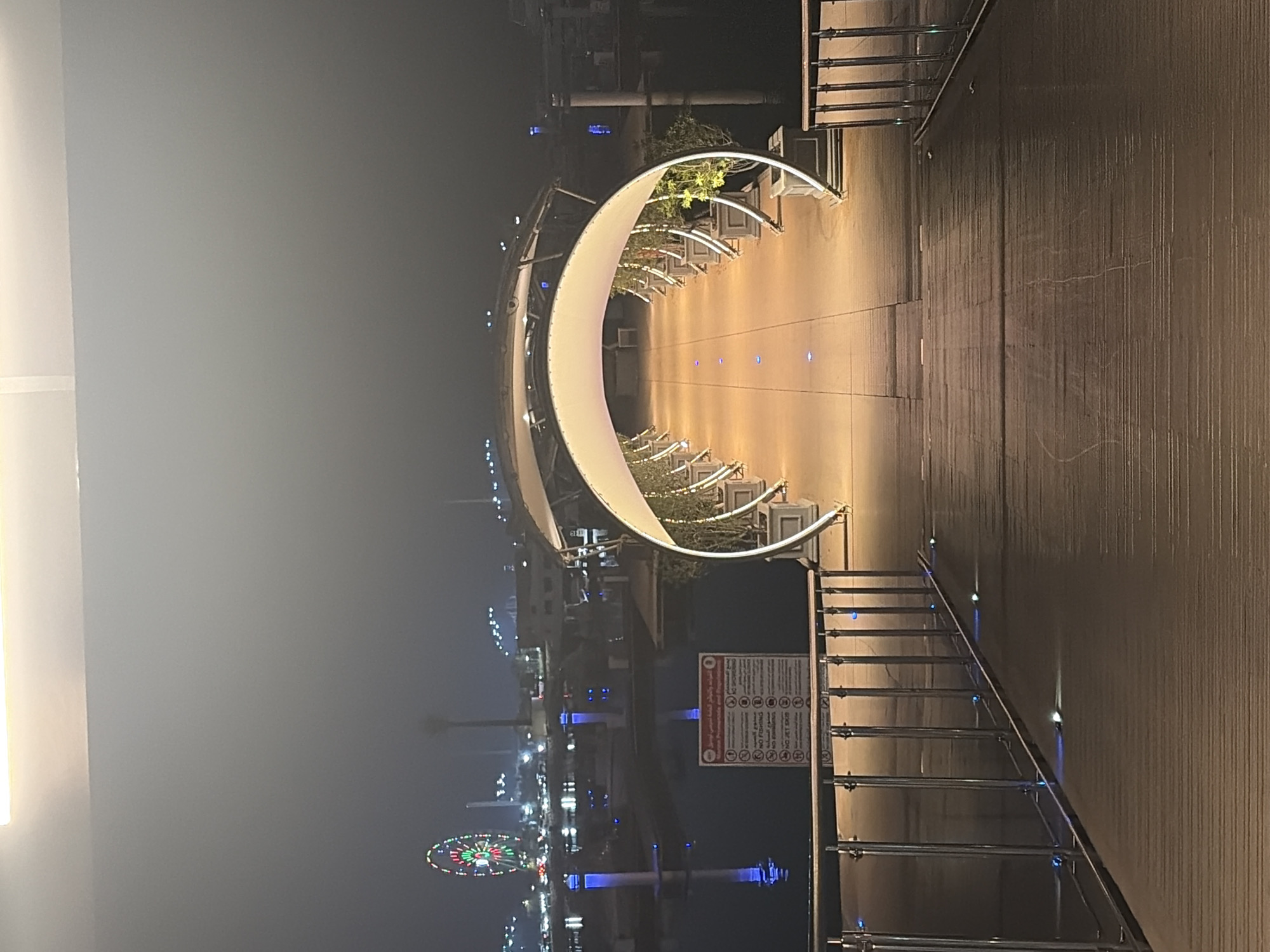 An illuminated pathway with a white arched canopy arches over a wooden walkway at night. The pathway is lined with potted plants. In the background, a Ferris wheel with colorful lights is visible, along with other city lights and reflections on dark water. A sign with regulations in Arabic and English is posted near the water. The scene appears to be in a marina or waterfront area.
