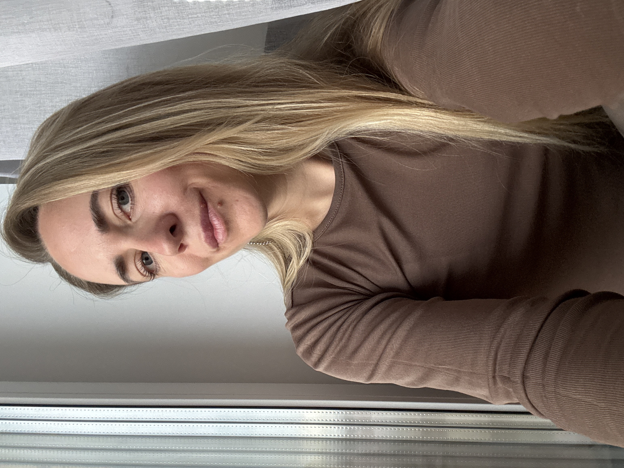 A portrait-oriented image, currently displayed on its side, features a young woman with long, wavy blonde hair and light-colored eyes, possibly blue or grey. She is depicted indoors, positioned in front of or next to a window, capturing a selfie or a close-up shot.
The woman is wearing a fitted, long-sleeved top in a warm, solid brown color, which appears to have a subtle ribbed or textured fabric. Her head is tilted gently, and she looks upwards and slightly to the viewer's right, displaying a faint, relaxed smile. Her expression conveys a sense of calm and pleasantness.
The background primarily consists of a window area. Towards the bottom of the frame, horizontal window slats or blinds are visible, suggesting an architectural feature. Above this, a light grey or white curtain or blind partially covers the upper portion of the window. The presence of natural light indicates it is daytime, although no specific time of day or weather conditions can be determined.
There is no visible text within the image. The provided location context for this scene is Chaikovsky, Russia.