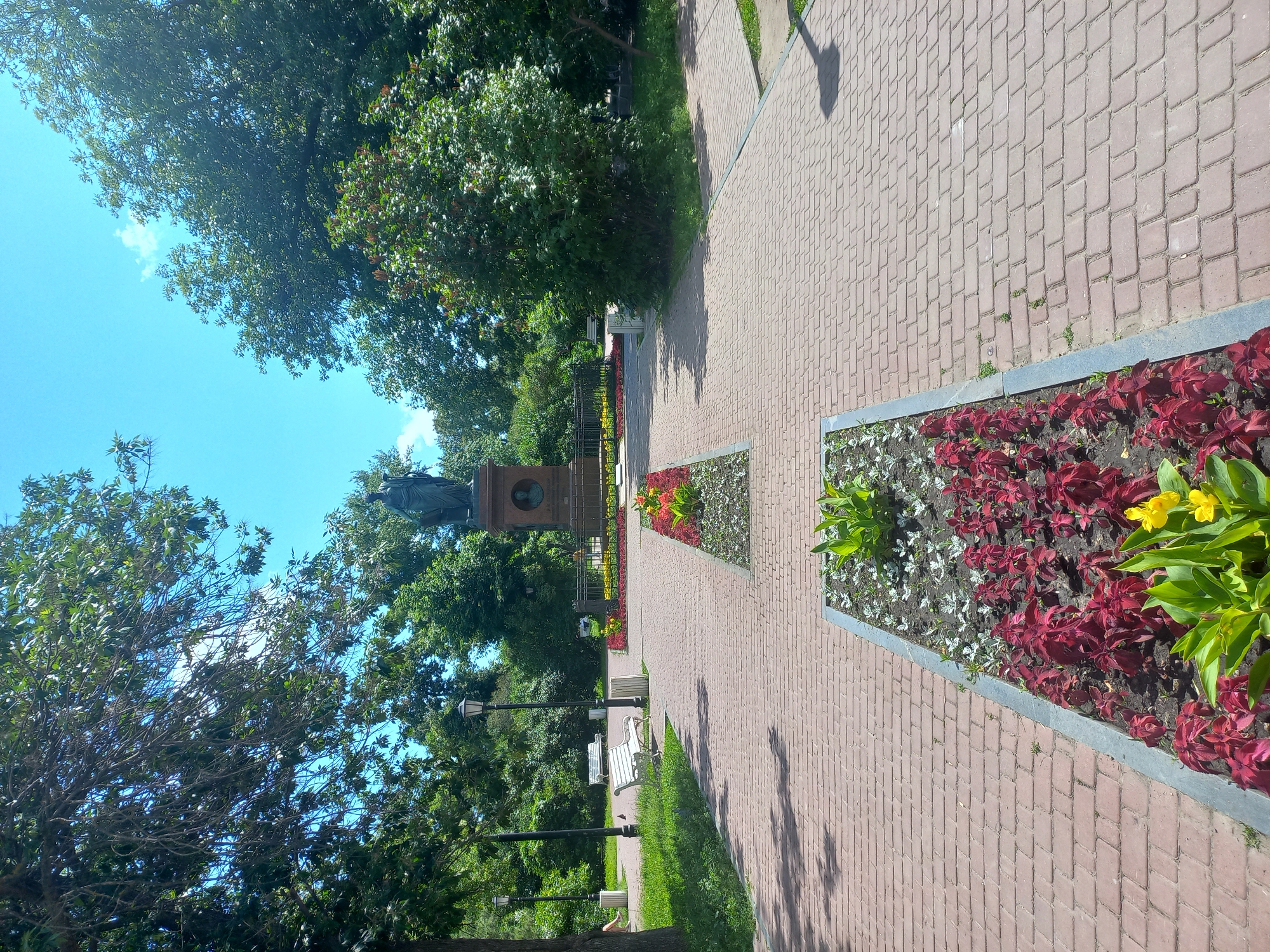 The image shows a park in Ulyanovsk, Russia, with a paved walkway leading towards a monument. The walkway is made of red bricks laid in a basket weave pattern. There are flowerbeds on either side of the walkway, with red and yellow flowers. In the background, there is a statue on a pedestal, surrounded by a black metal fence. There are trees and benches in the park, and the sky is blue with some clouds. The scene appears to be peaceful and well-maintained.
