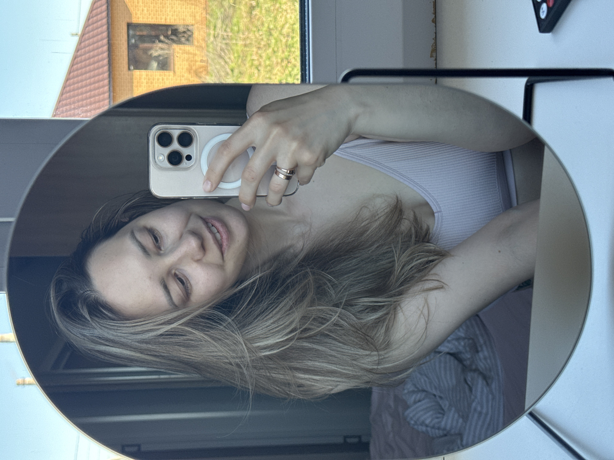 A woman with long, highlighted hair is taking a selfie in an oval mirror. She is holding a gold-colored iPhone with a MagSafe ring on the back. The woman is wearing a light-colored tank top. Reflected in the mirror are elements of a window with a view of a building and greenery outside. A white wall and part of a black object appear next to the mirror. A striped pattern can be seen behind the woman, possibly a bed cover. Based on information from the user, the location is Chaikovsky, Russia.