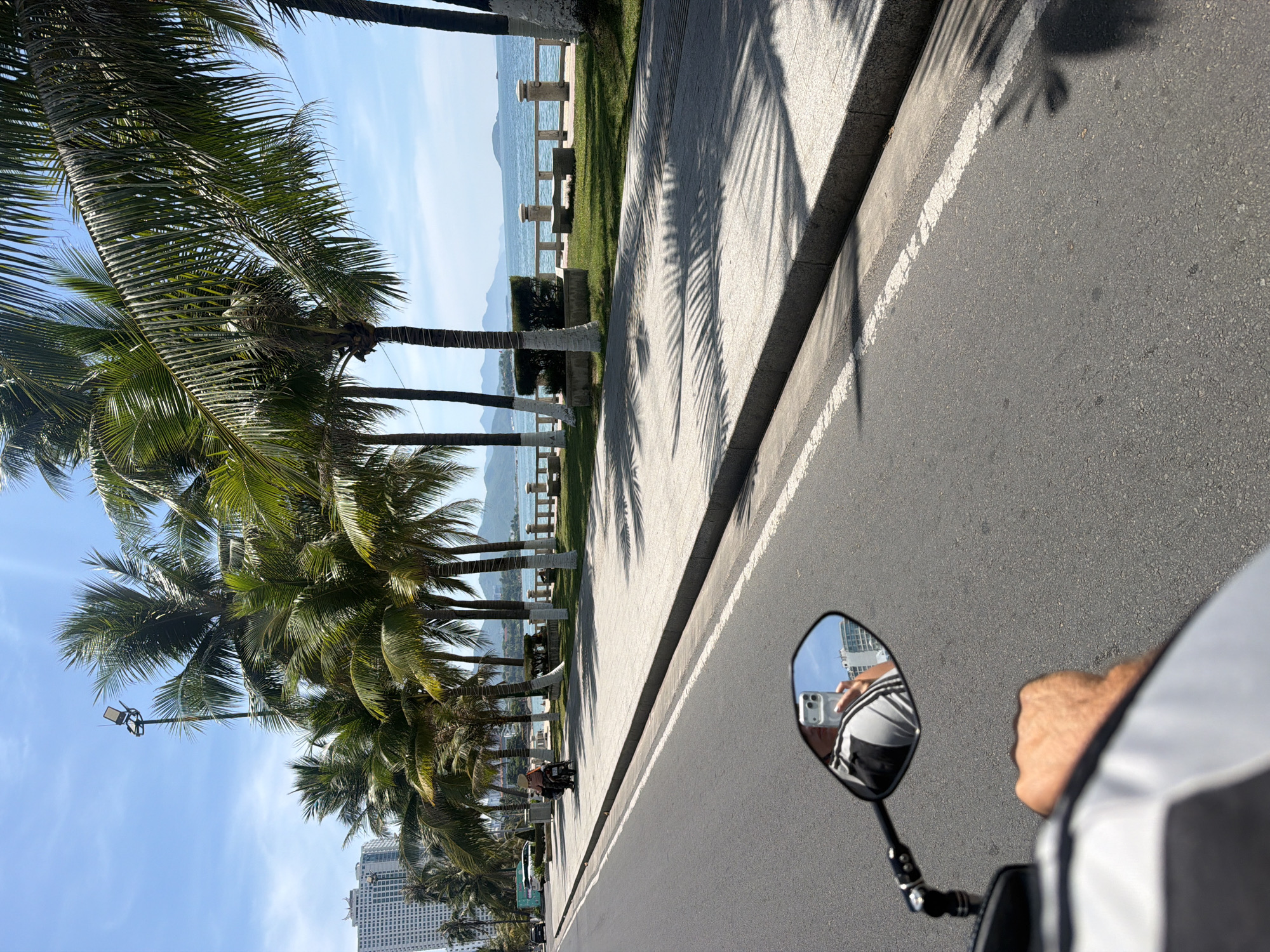 The image depicts a sunny day in Nha Trang Ward, Vietnam, with a perspective from a moving vehicle, likely a motorcycle, looking forward along a coastal road. In the foreground, a rider's hand grips the handlebar, and a motorcycle mirror reflects the rider taking a selfie with a smartphone. Beyond the mirror, the road is visible, bordered by a paved walkway lined with palm trees.
The walkway leads to a grassy area with benches, a railing, and the ocean beyond. In the distance, mountains are visible under a clear blue sky with scattered clouds. A tall building is also seen in the background on the left side of the image. The scene suggests a tropical coastal city environment.