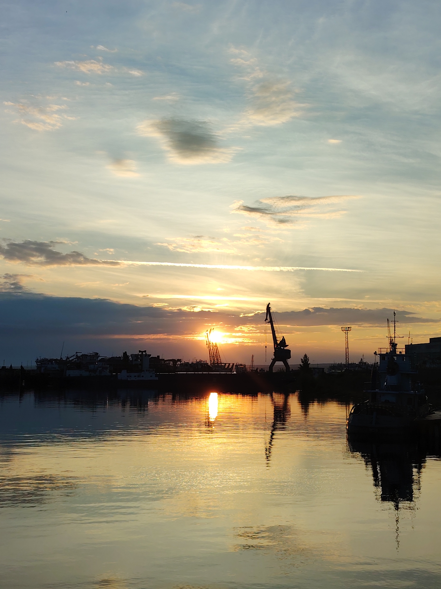 This is a vertical, full-shot photograph taken at a harbor in Chaikovsky, Russia, during a sunset. The sky is a gradient of pale blue at the top, transitioning to soft oranges and yellows near the horizon, with scattered clouds. The sun, a bright, diffused orb, is positioned low in the sky, casting a warm glow and creating a strong reflection on the calm water.
In the middle ground, a line of silhouetted ships and industrial cranes are visible against the sunset. The water in the foreground is smooth and reflects the sky, clouds, and the outlines of the ships and cranes. No people are visible in the image. The overall atmosphere is serene and tranquil, emphasizing the beauty of the natural light over the industrial landscape.