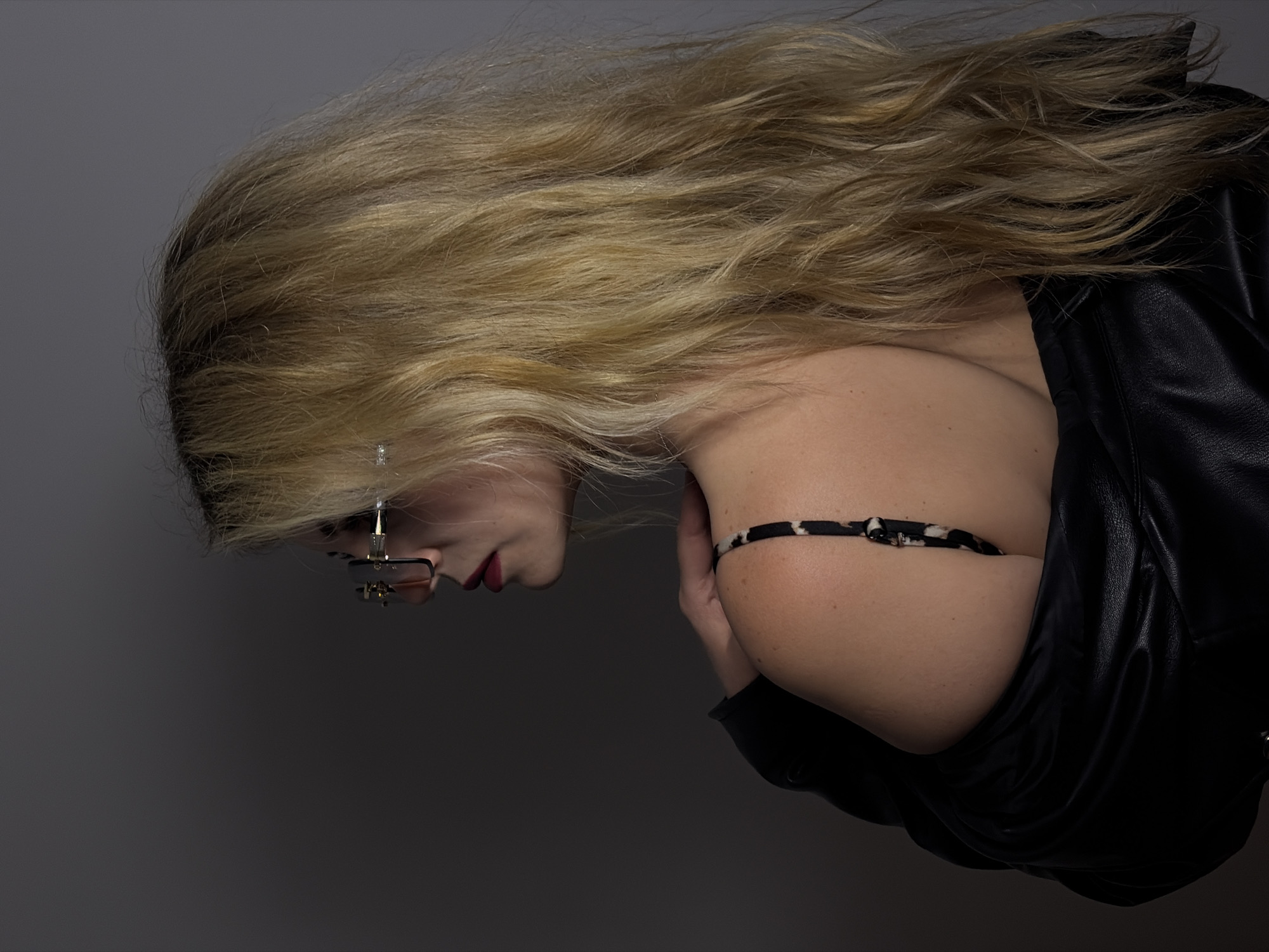A medium shot depicts a fair-skinned woman with long, wavy, blonde hair. She is wearing black-rimmed glasses with a multi-bridged nose piece. Her lips are colored red. She is wearing a black leather jacket and a black and white spotted strap. The background is a neutral grey. There is no evidence in the image to suggest it was taken in Budva, Montenegro.