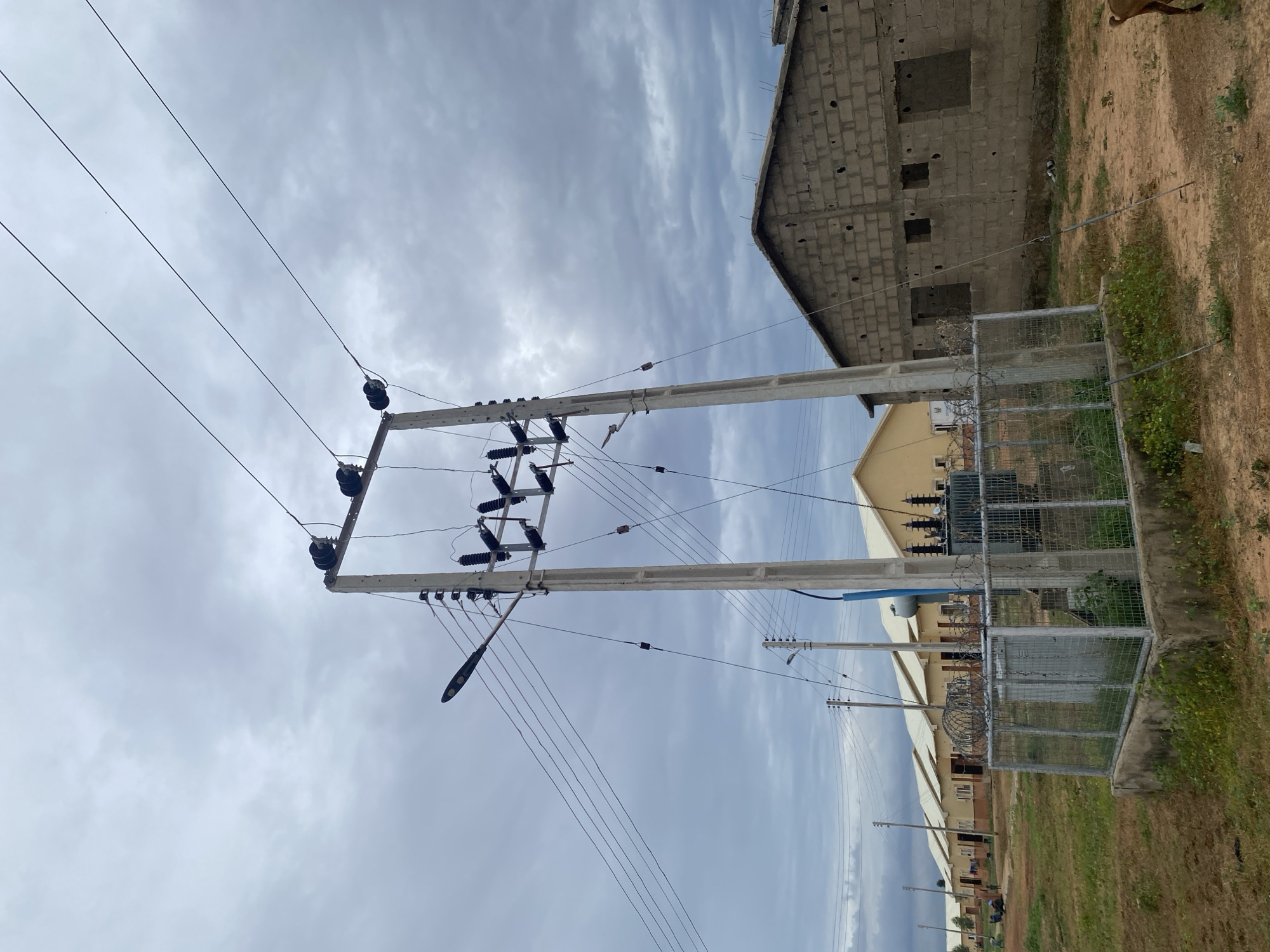 This media file captures an outdoor scene dominated by overhead power lines and utility poles. The power lines stretch across the frame, supported by concrete poles equipped with insulators. A street light is attached to one of the poles. In the background, a building under construction, made of cinder blocks with open window frames, stands against a cloudy sky. Adjacent to this, another building with a yellow facade and a fenced enclosure featuring barbed wire can be seen. The ground is a mix of dry earth and sparse vegetation. The sky is overcast, suggesting a cloudy or possibly rainy day. The scene appears to be in Dumnga, Nigeria. There are no discernible people or animals actively engaged in any activity. The overall mood is neutral, focusing on the infrastructure and environment.