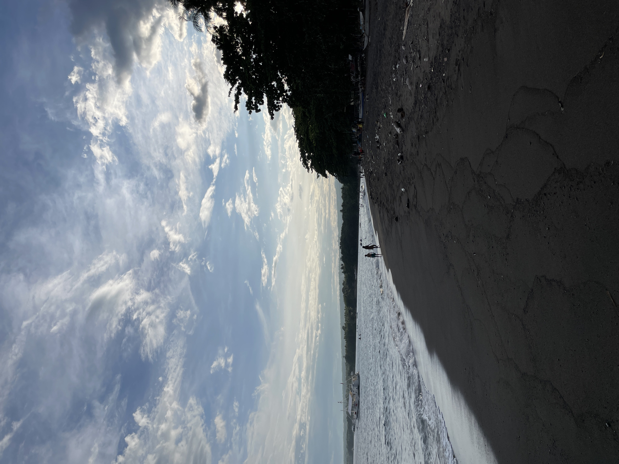 The image depicts a beach scene in Dauin, Philippines. The foreground shows dark, fine sand with some debris scattered on its surface, including plastic bottles, wood fragments, and what appear to be coconuts. The sand is textured with subtle patterns formed by the receding tide. To the left, the ocean waves gently roll onto the shore, creating a foamy white edge. The water is a deep grey-blue. In the mid-ground, a large white boat with multiple levels and flags is anchored offshore. Several people are visible in the water and walking along the shoreline in the distance.
The background features a lush, dark green treeline that runs along the coast. The trees are dense and appear to be tropical vegetation. The sky is a dramatic mix of blue and white, with scattered clouds that are illuminated by the sun, creating a sense of diffused light. The overall atmosphere suggests a late afternoon or early evening setting. The presence of the boat and people indicates recreational activity or local life on the beach.